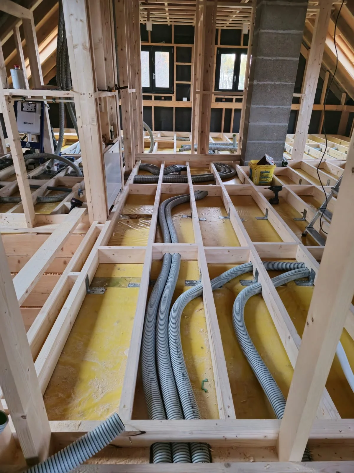 The interior of a house under construction showing the framework with wooden studs and electrical wiring and conduits installed in the floor and walls, with two windows visible in the background.