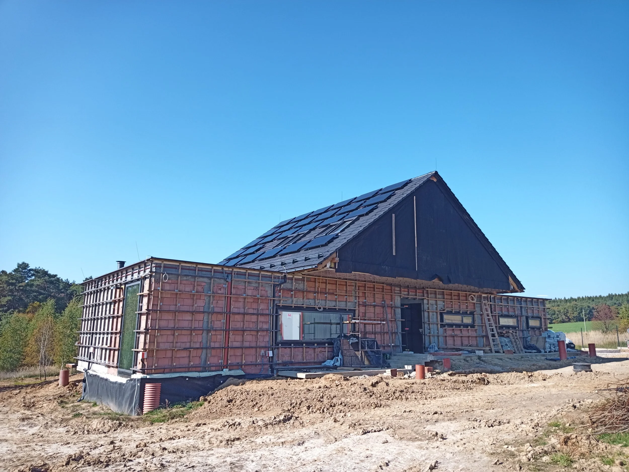 A house under construction with a steep roof covered in solar panels, brick walls, and construction materials around the site, set against a backdrop of green trees and a clear blue sky.