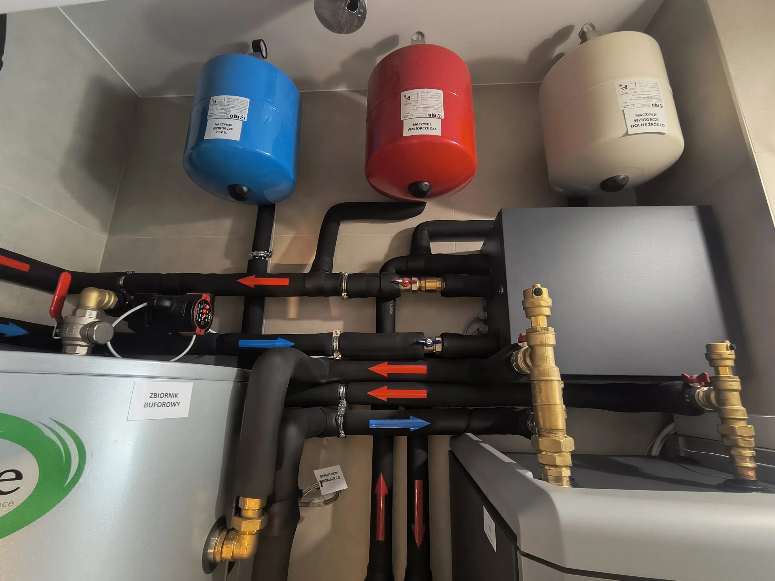 A plumbing system with pipes, valves, and tanks, including a blue, red, and white container labeled in Polish, with various pipes and fittings connected, and labels indicating their purpose.