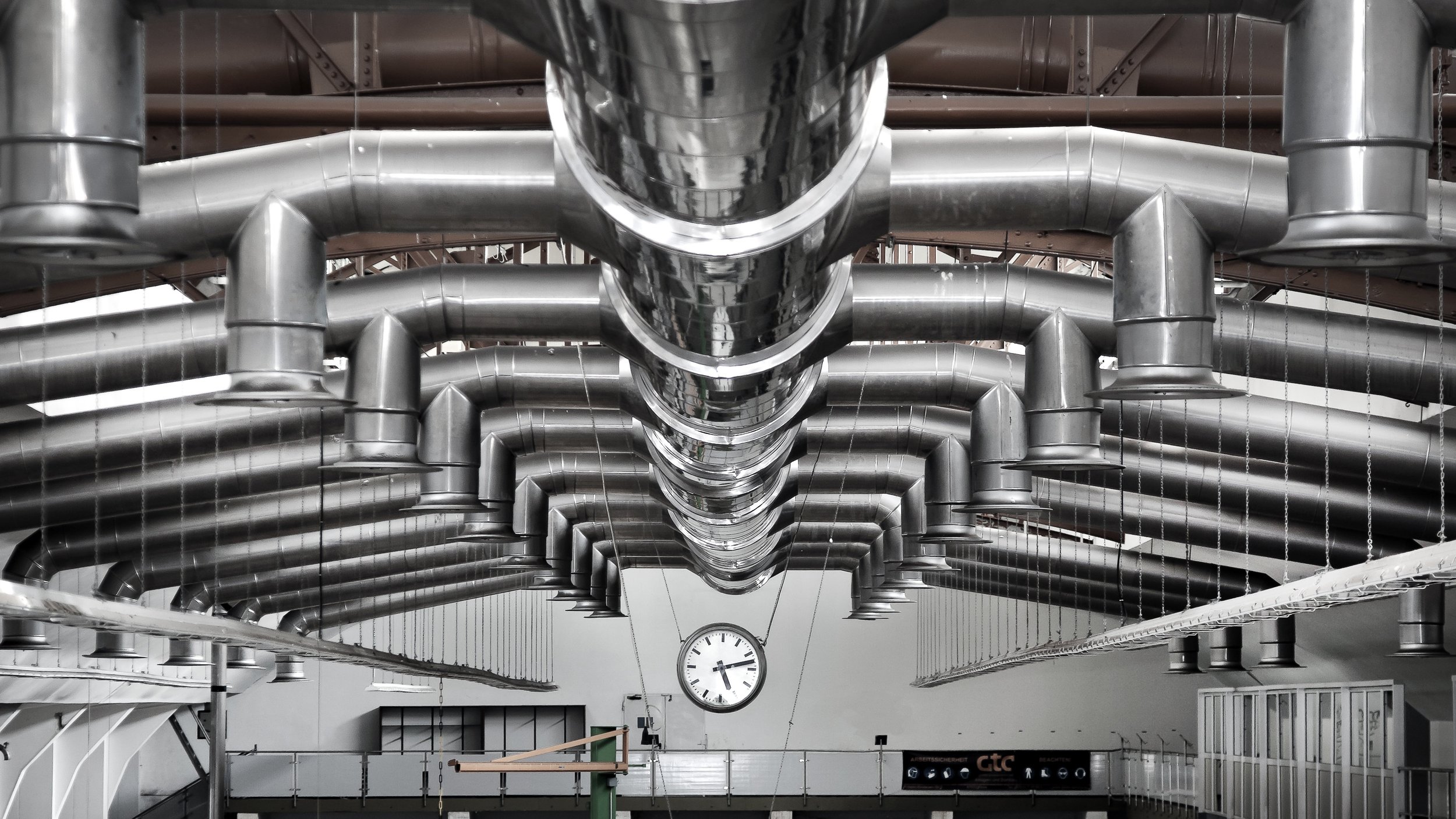 A ceiling with multiple large, silver metallic air ducts and pipes arranged in parallel, with a wall-mounted clock displaying 3:24 in the background.