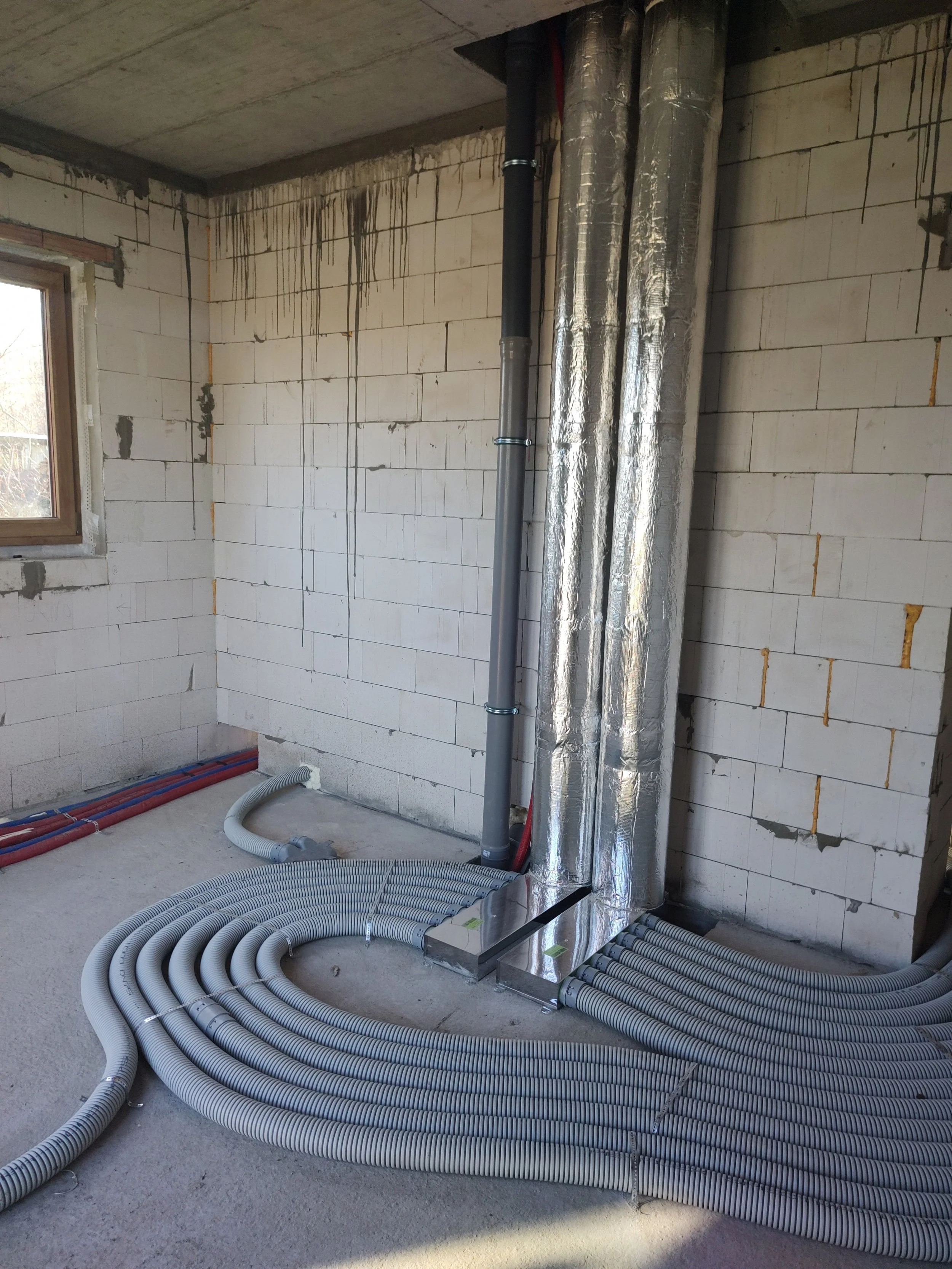 Construction site with exposed pipes and ducts, unfinished brick walls, and a window, indicating ongoing building or renovation work.