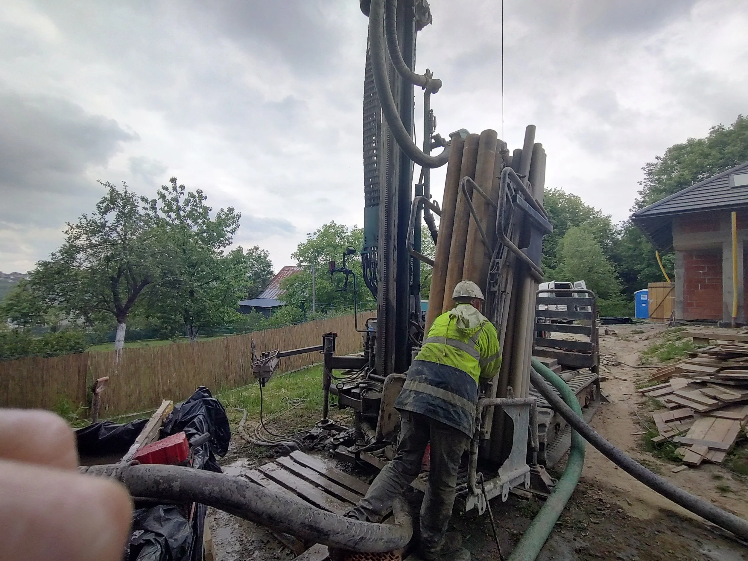 Construction worker wearing a safety helmet and neon yellow jacket operating heavy machinery on a construction site under a cloudy sky, with trees, a fence, and a house in the background.