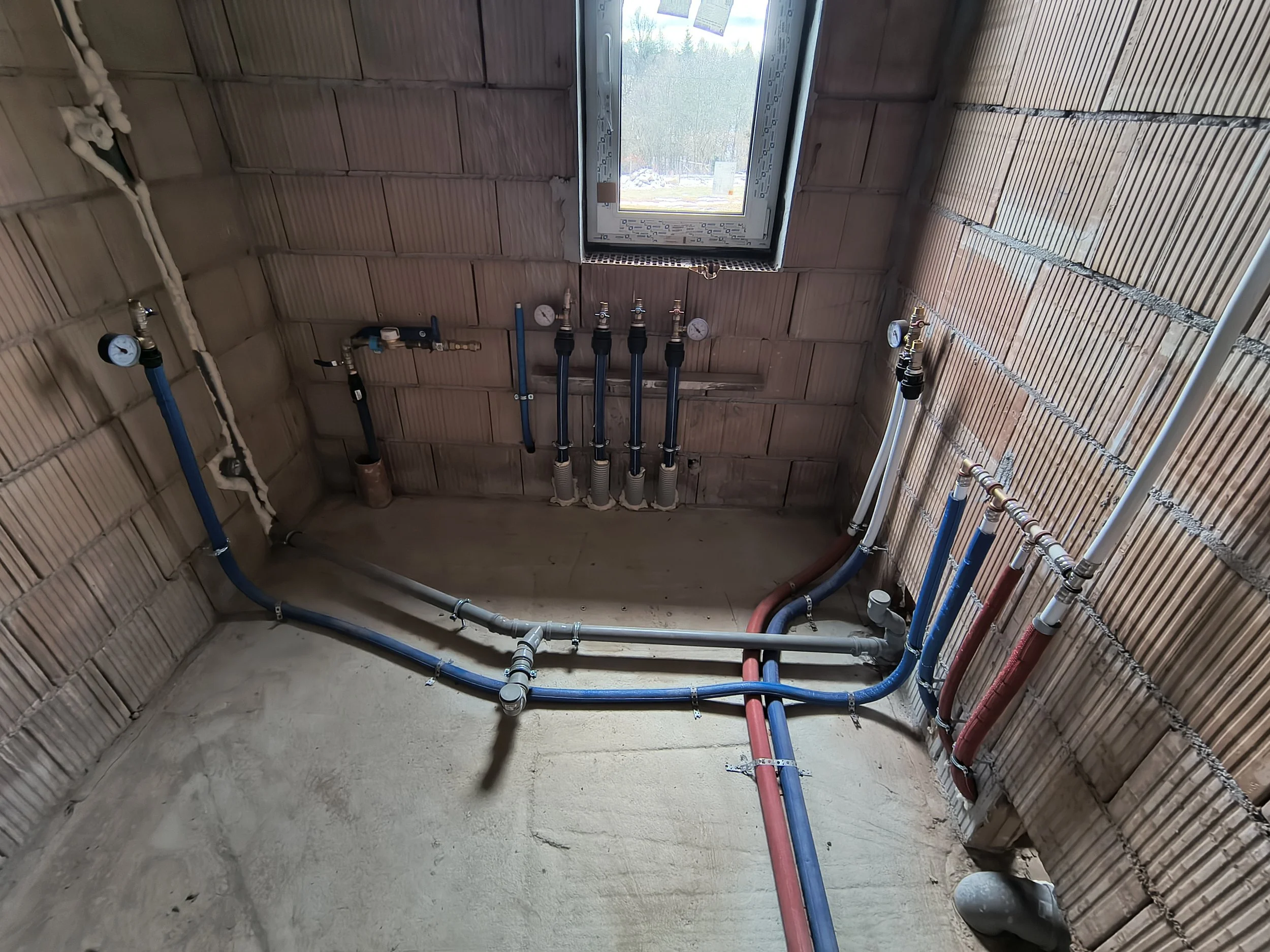 Interior view of a room under construction with plumbing pipes installed in the wall and ceiling, an open window showing outdoor scenery, and unfinished brick walls.
