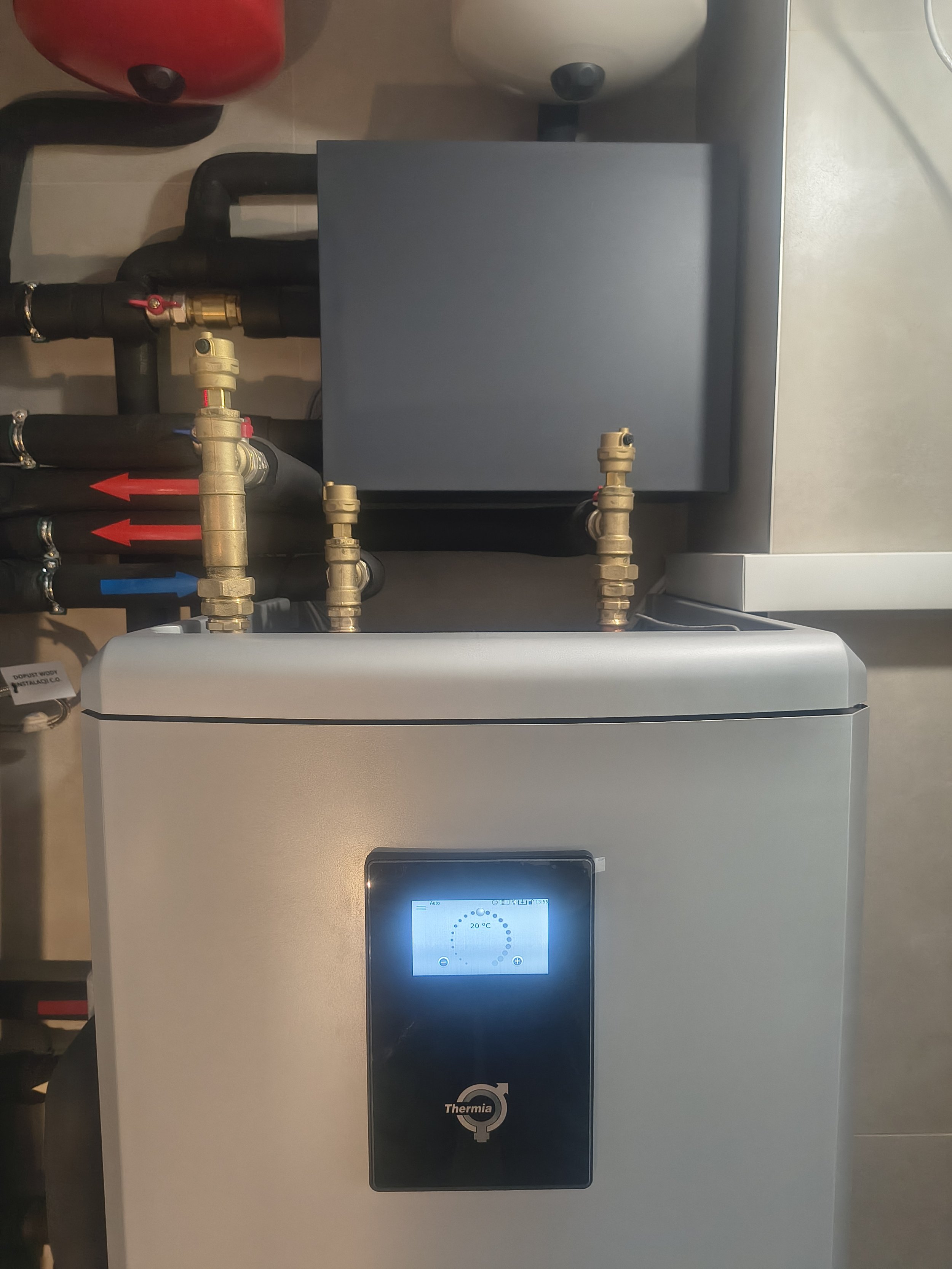 A modern heating or water system with a digital display, branded Thermia, connected to black pipes with gauges and valves, situated against a light-colored wall.