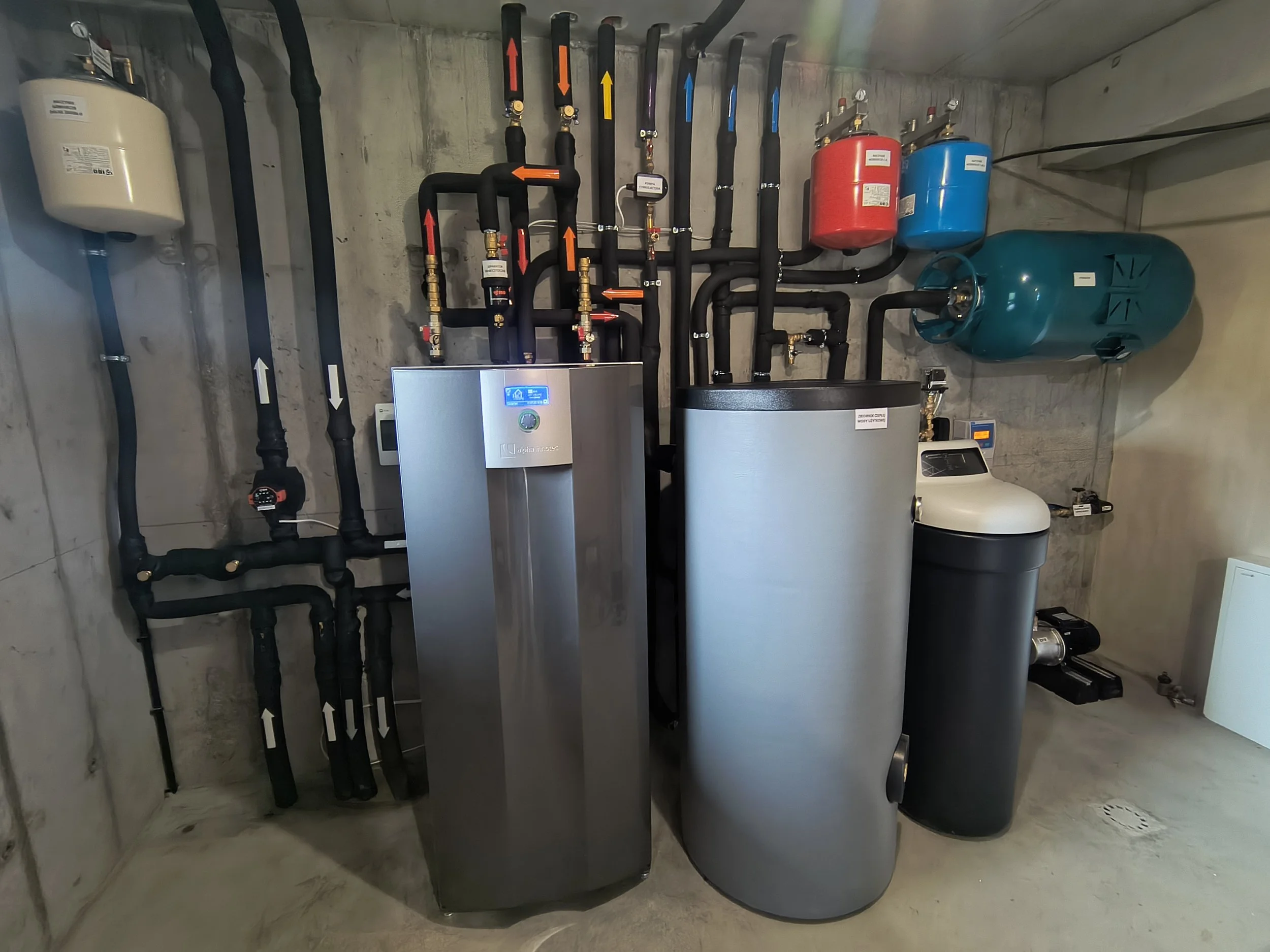 The image shows a mechanical room with various pipes and tanks. There are black pipes with colored arrows, a digital control panel, and two large water tanks, one red and one blue. The room has a concrete wall and floor.