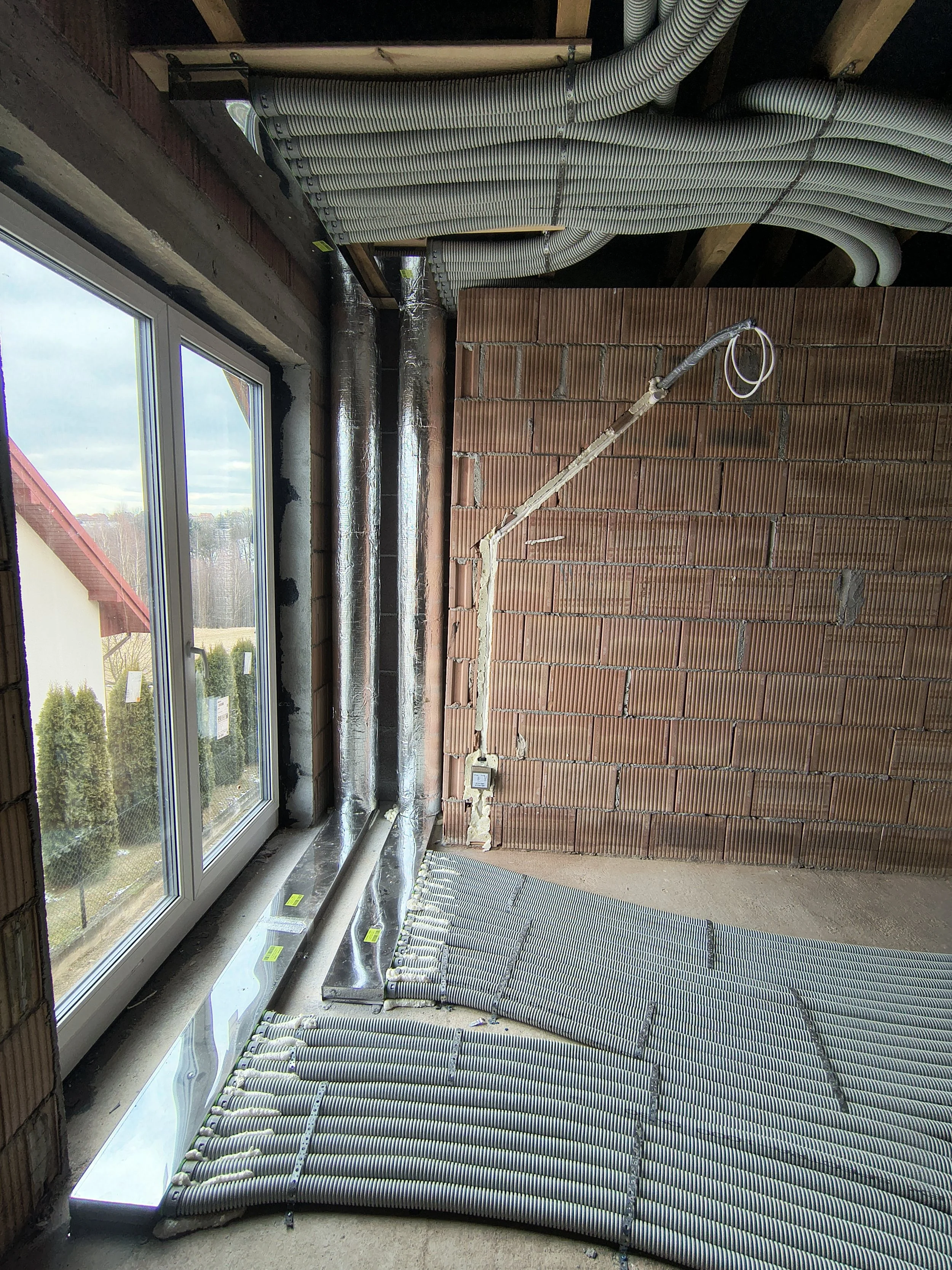 Interior of a building under construction with exposed brick wall, ceiling ducts, and a large window showing an outdoor view of trees and a cloudy sky.
