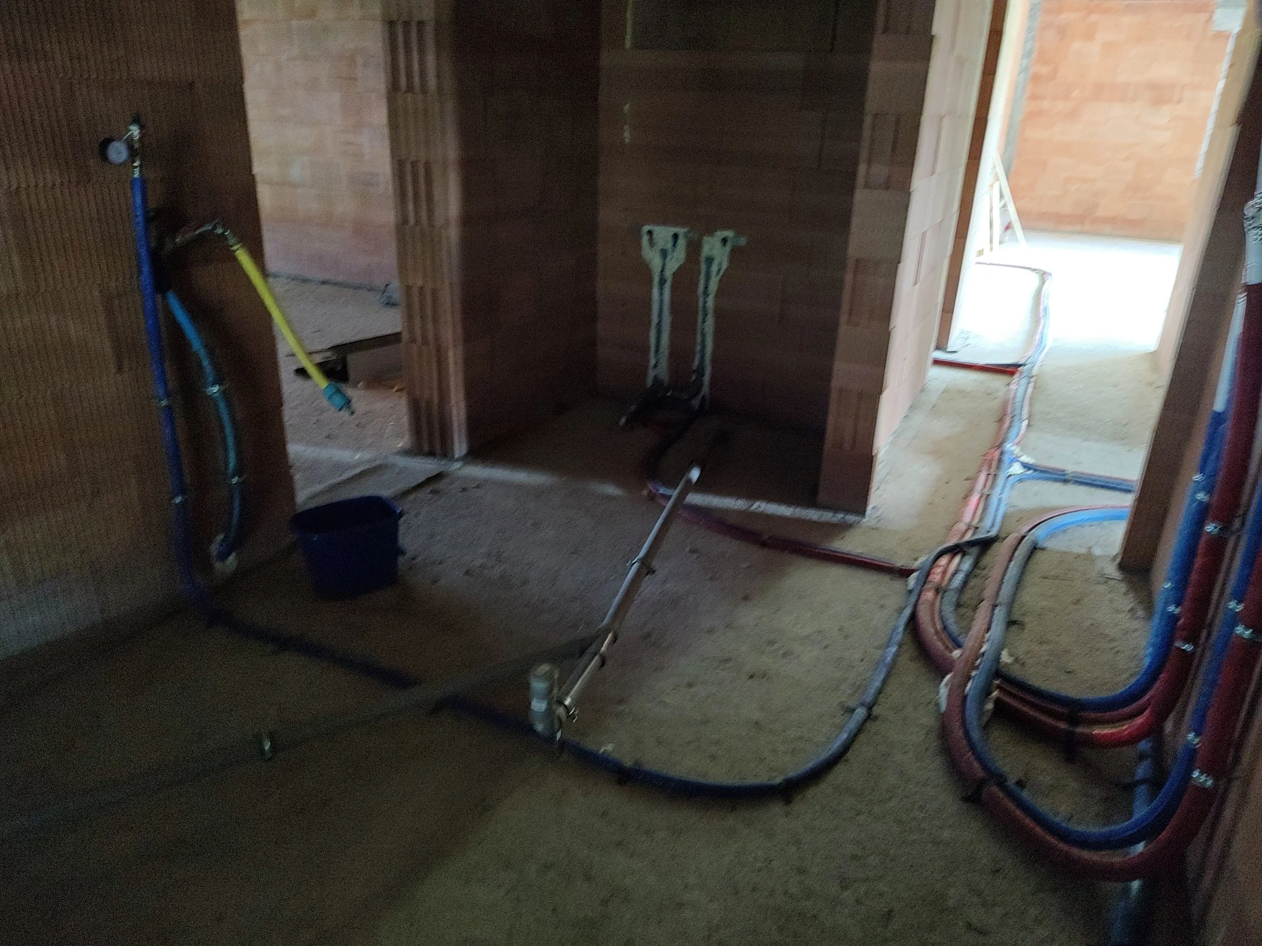 The image shows an unfinished room with brick walls and exposed electrical wiring. There are two wiggle sticks on a dark wooden wall, and a blue bucket on the floor. Also, there is a staircase leading to an upper level, with more electrical wiring vi