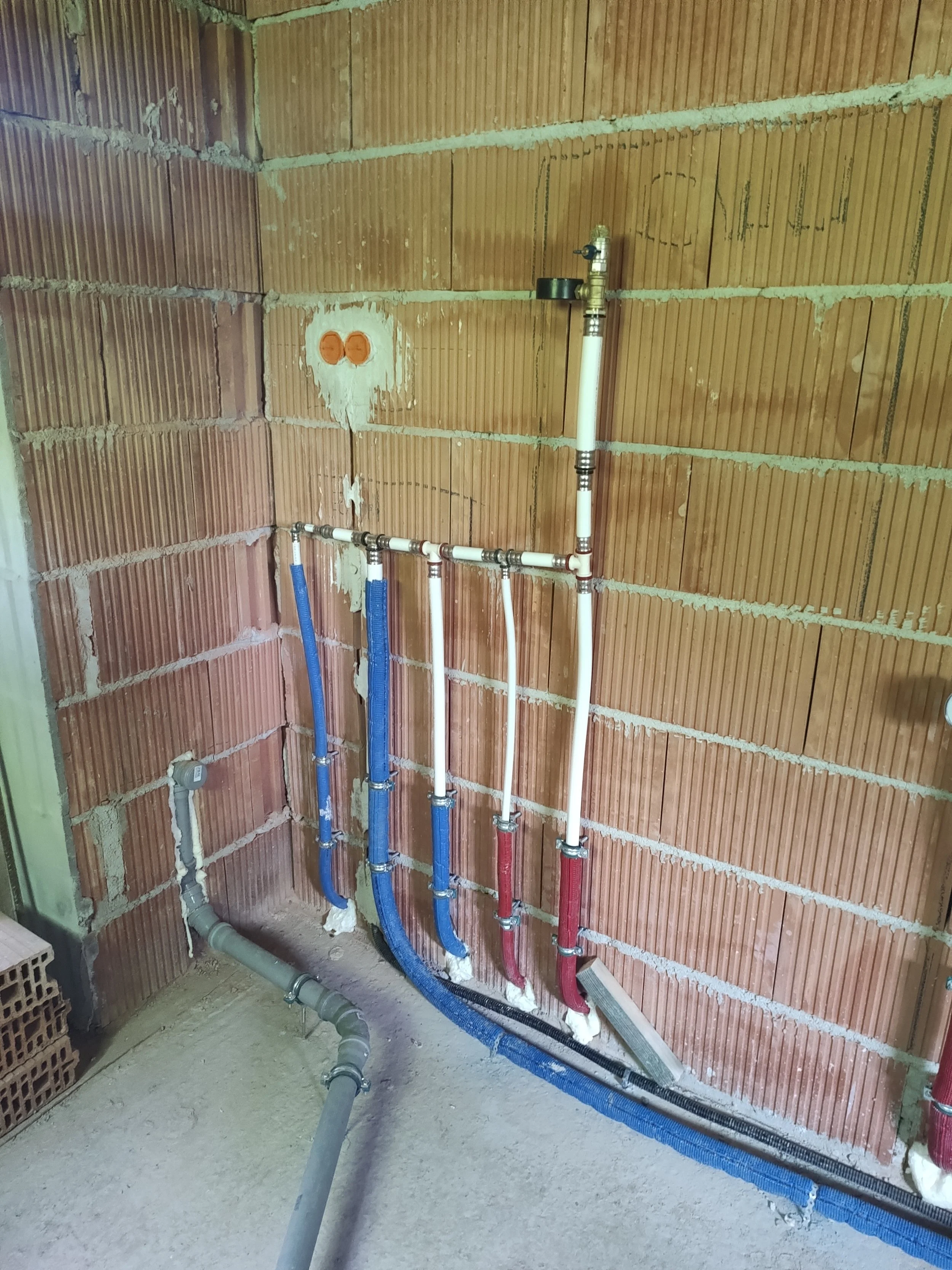 Plumbing pipes in a wall with visible brickwork, including white, blue, and red pipes, connected to a water valve.