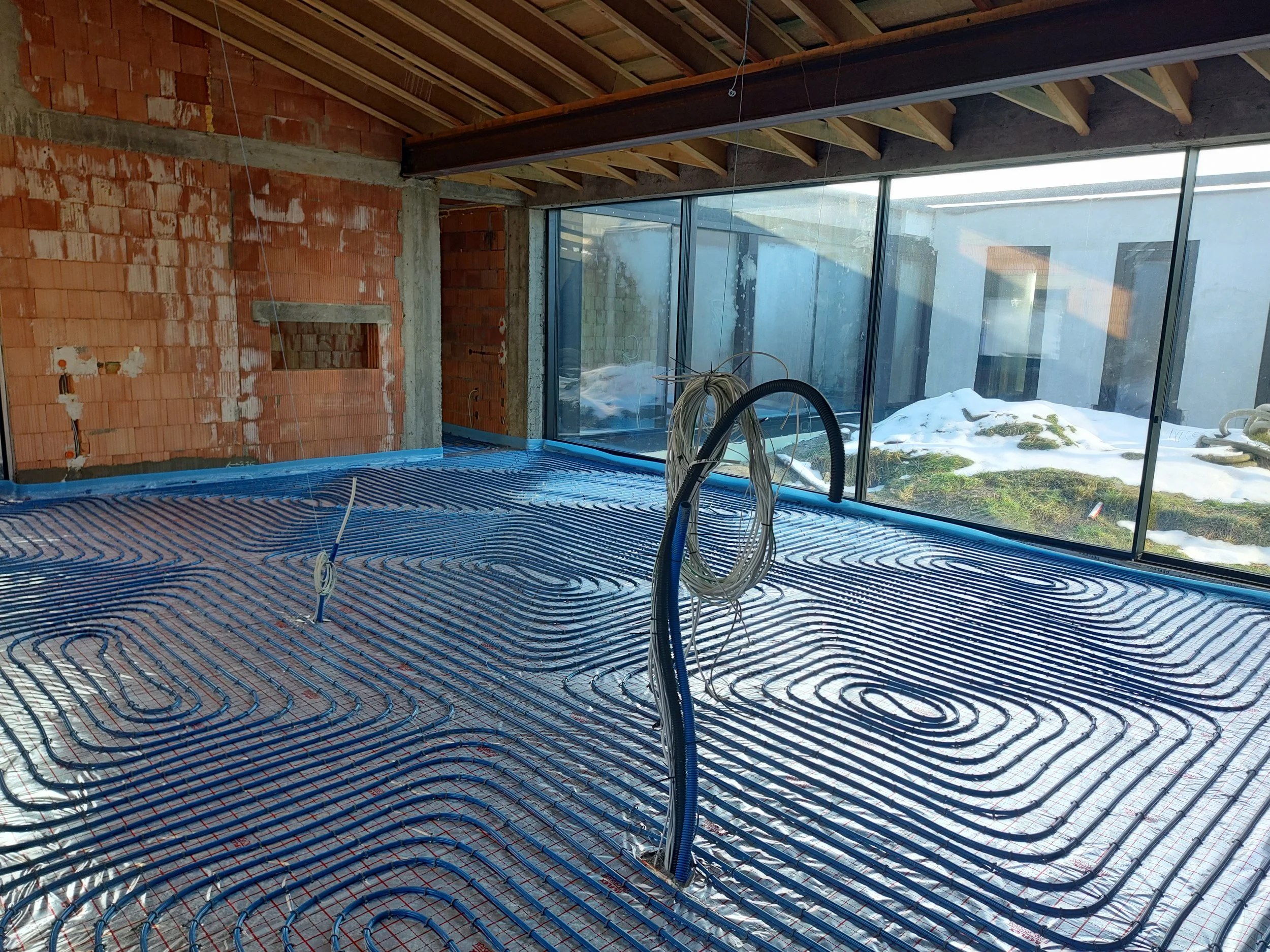 Under construction room with exposed brick walls and large glass windows. Radiant floor heating pipes are installed on the floor, with some electrical wiring hanging. Snow can be seen outside through the windows.