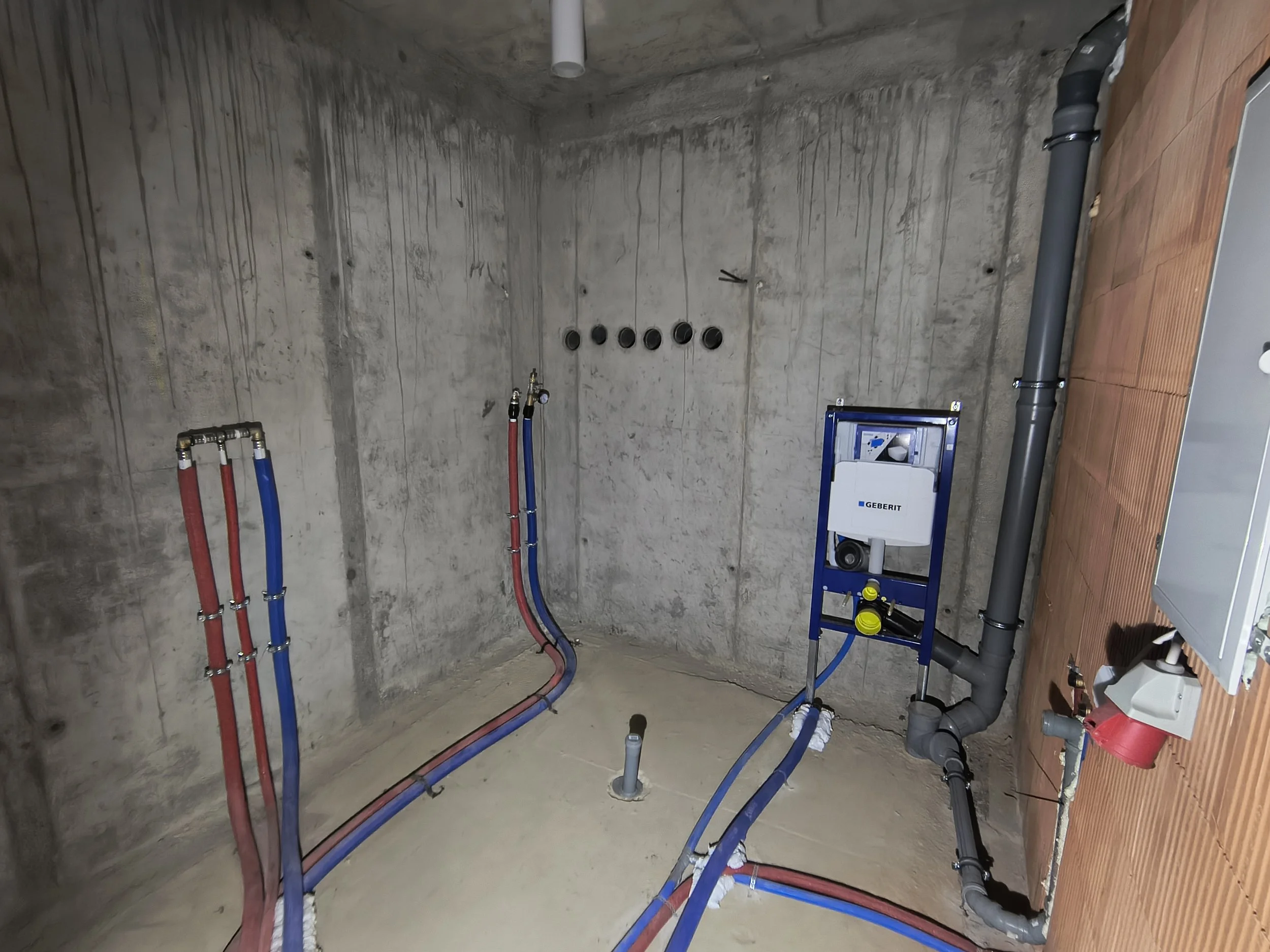 Construction space with exposed concrete walls and a network of red and blue pipes, along with various plumbing fixtures and a Geberit black piping system.
