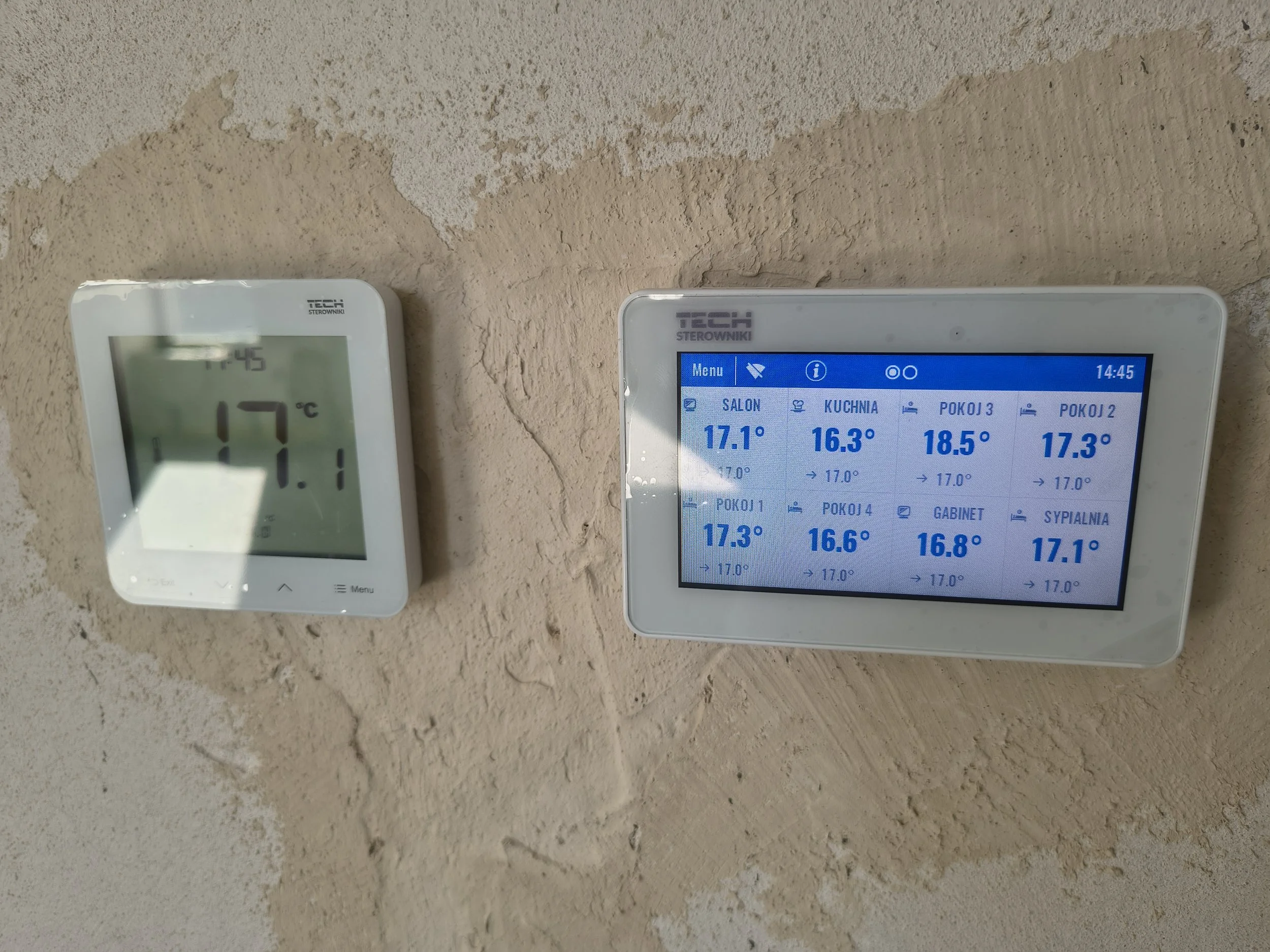 Two digital thermostats mounted on a textured beige wall, displaying times and temperature readings.
