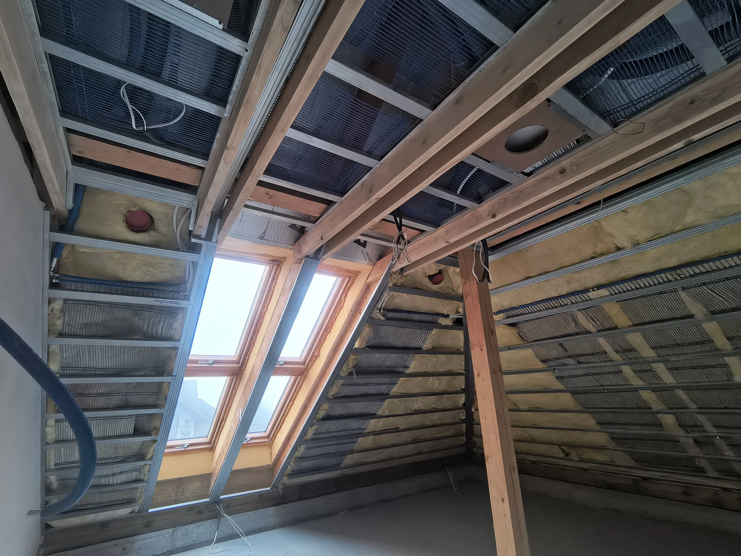 Interior view of a building under construction with exposed roof structure, solar panels, wiring, and insulation. A window is visible in the background.