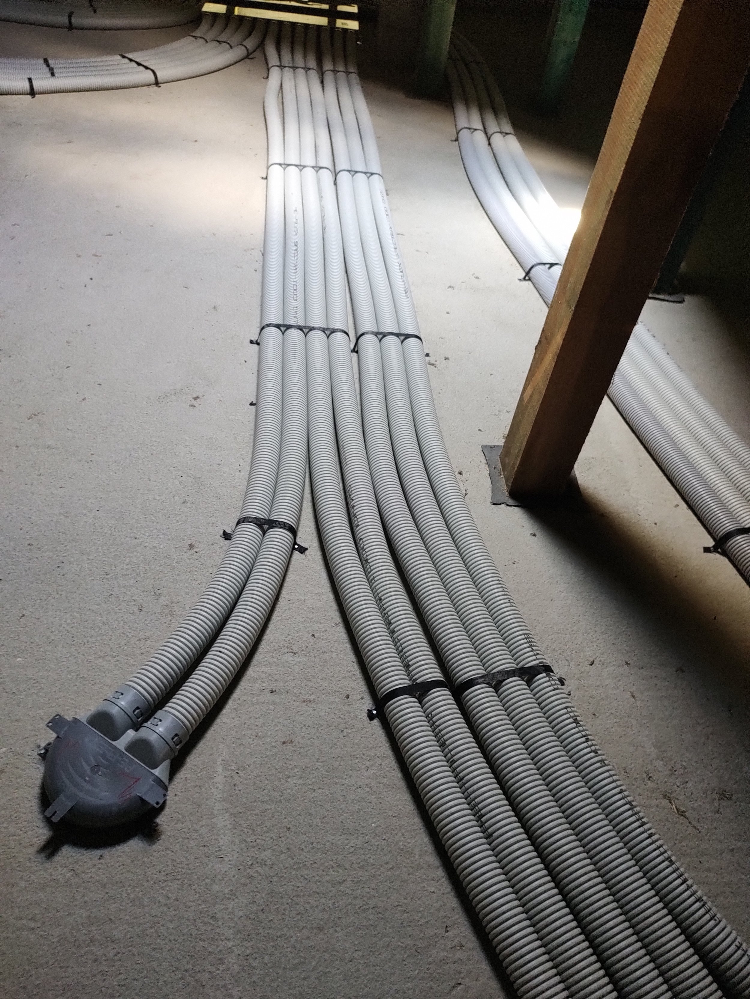 Several white flexible conduit pipes arranged on a concrete floor, connected to a vacuum cleaner, with some pipes secured with black ties, and a wooden support beam visible on the right side.