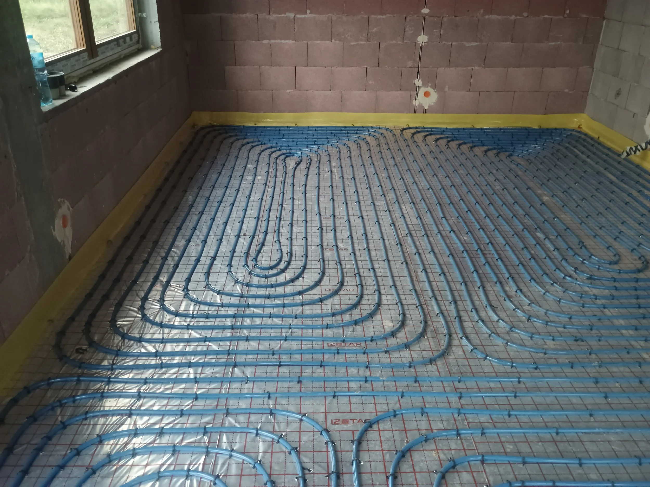 Underfloor radiant heating system installation in progress inside a room with unfinished brick walls. Blue PEX pipes are laid out in loops on a reflective insulation layer, with a window on the left wall showing an outdoor view.