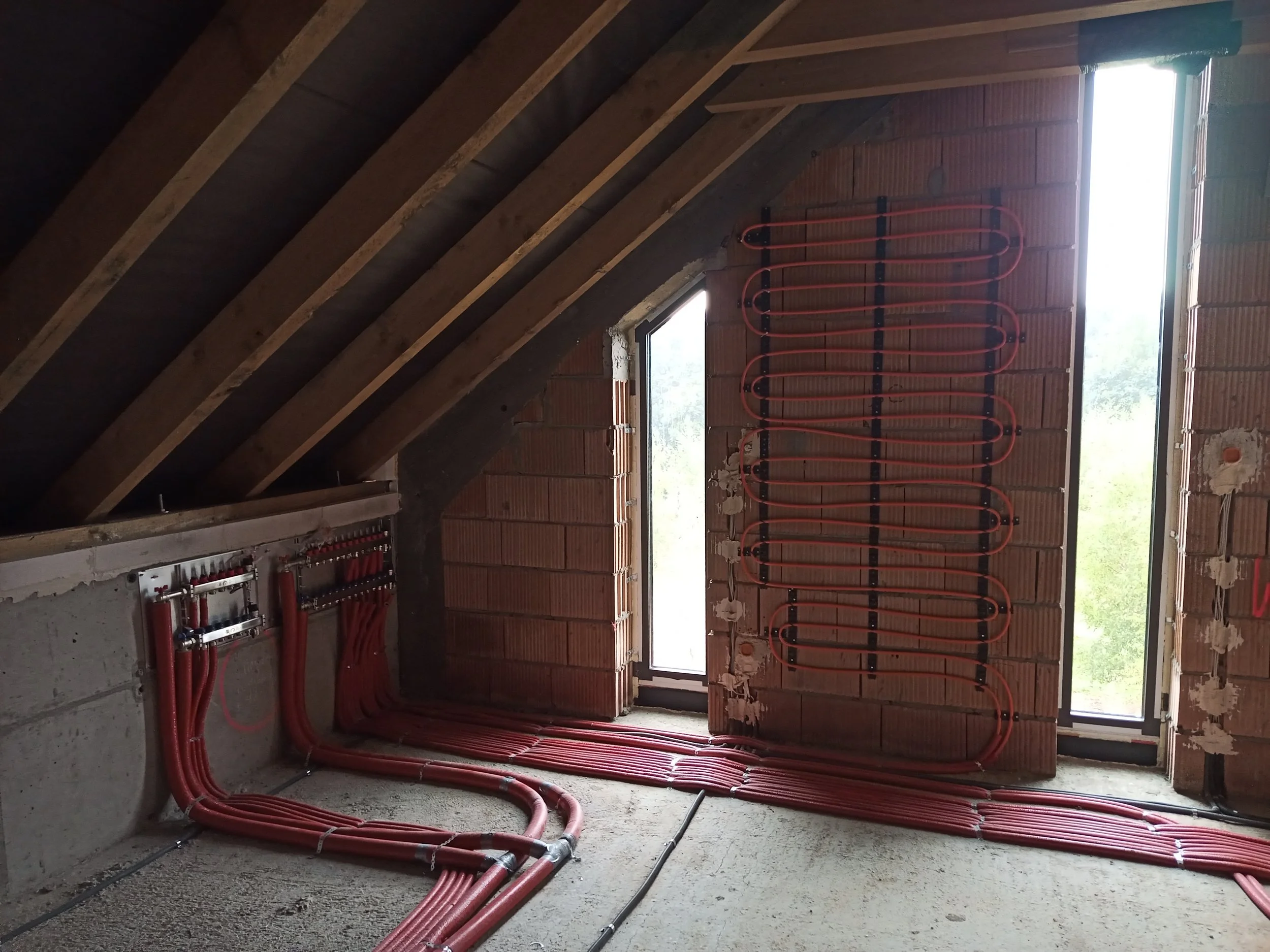 Interior of a building under construction with red piping and plumbing installed on the walls and floors, and large windows letting in natural light.