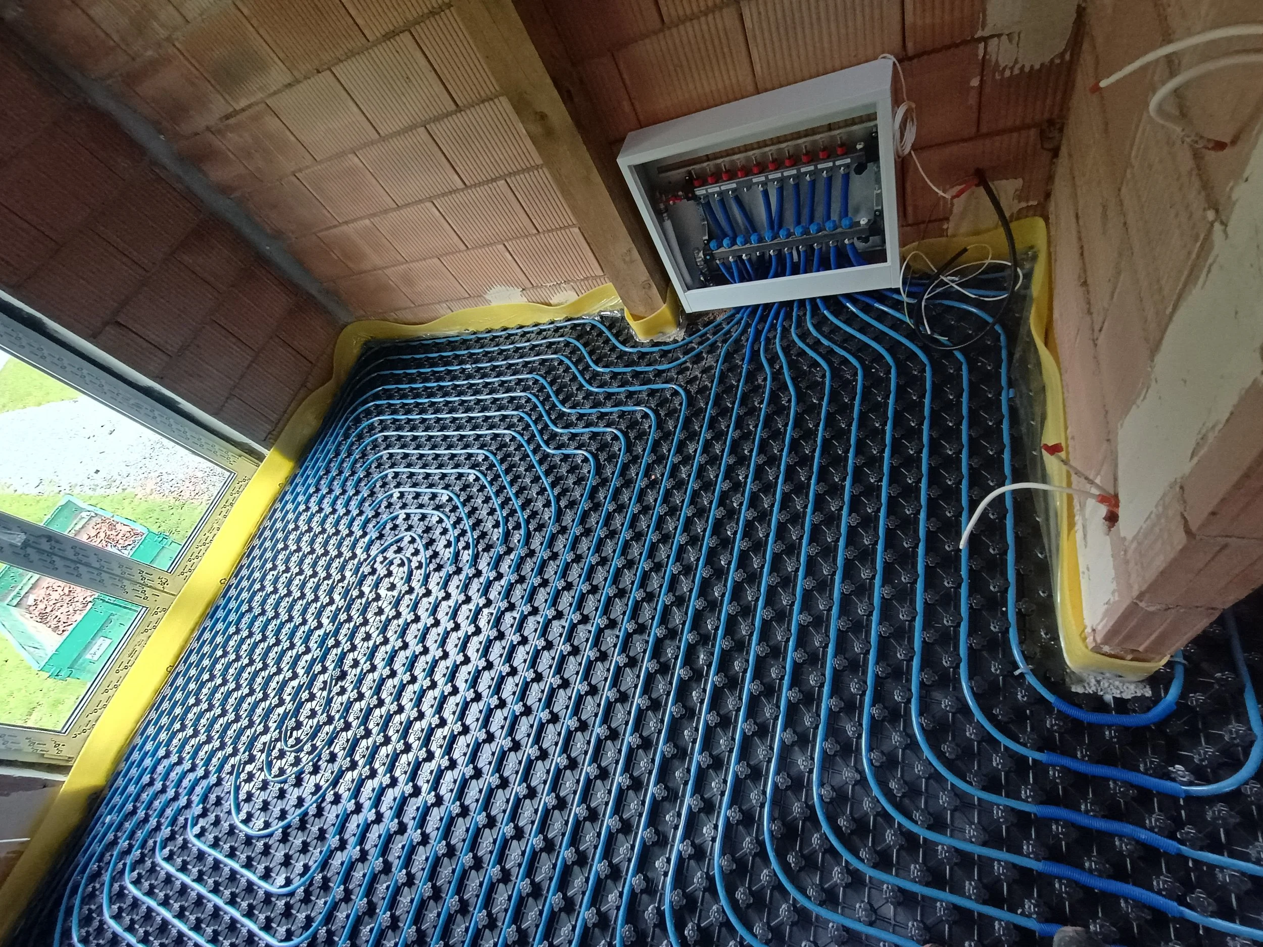 Installation of radiant floor heating system with blue PEX tubing in a room with brick walls and a window.