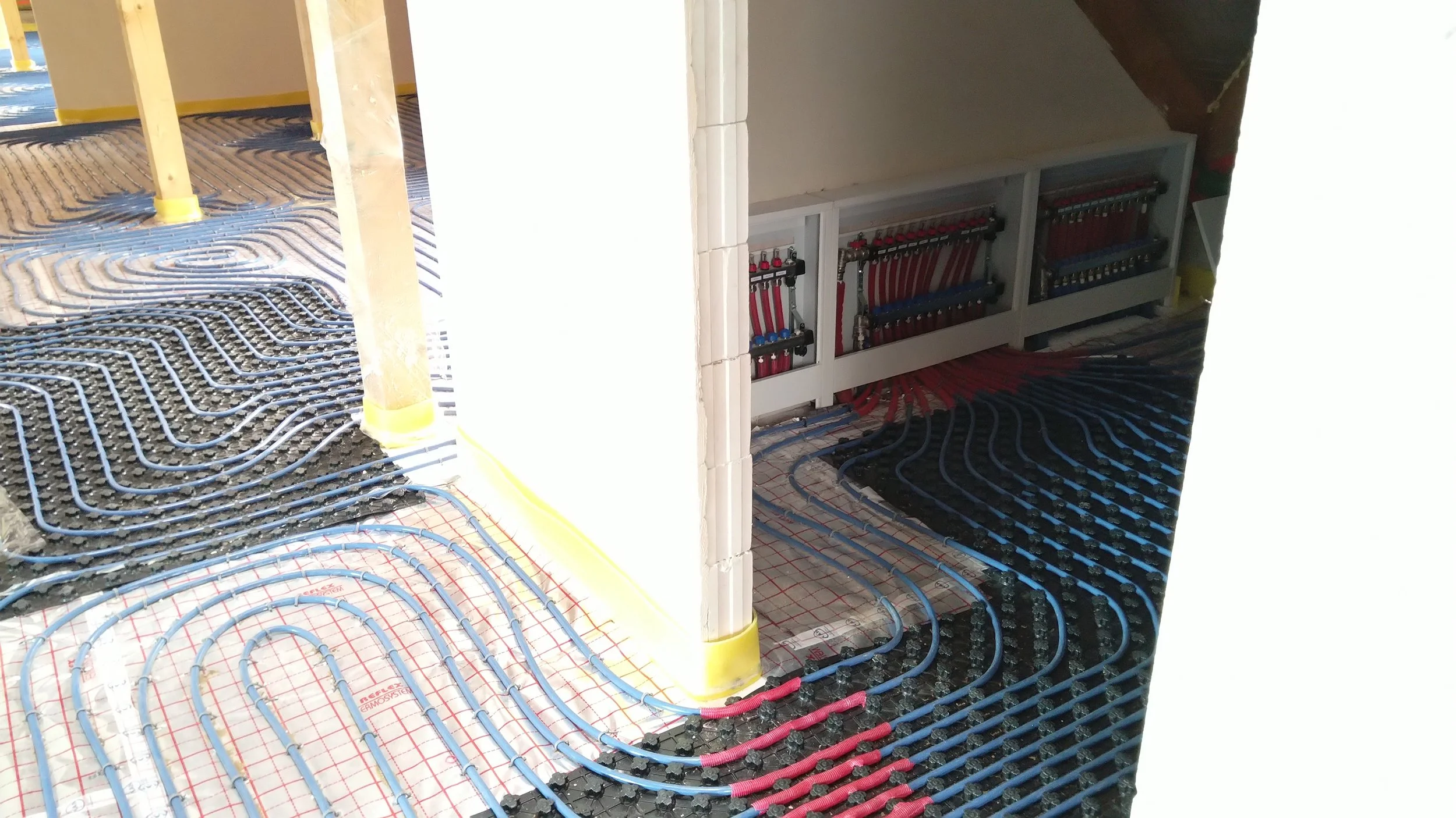 Underfloor heating system under construction, with blue and red PEX tubing laid in a pattern over insulation and a black foam board, near white walls and wooden support beams.