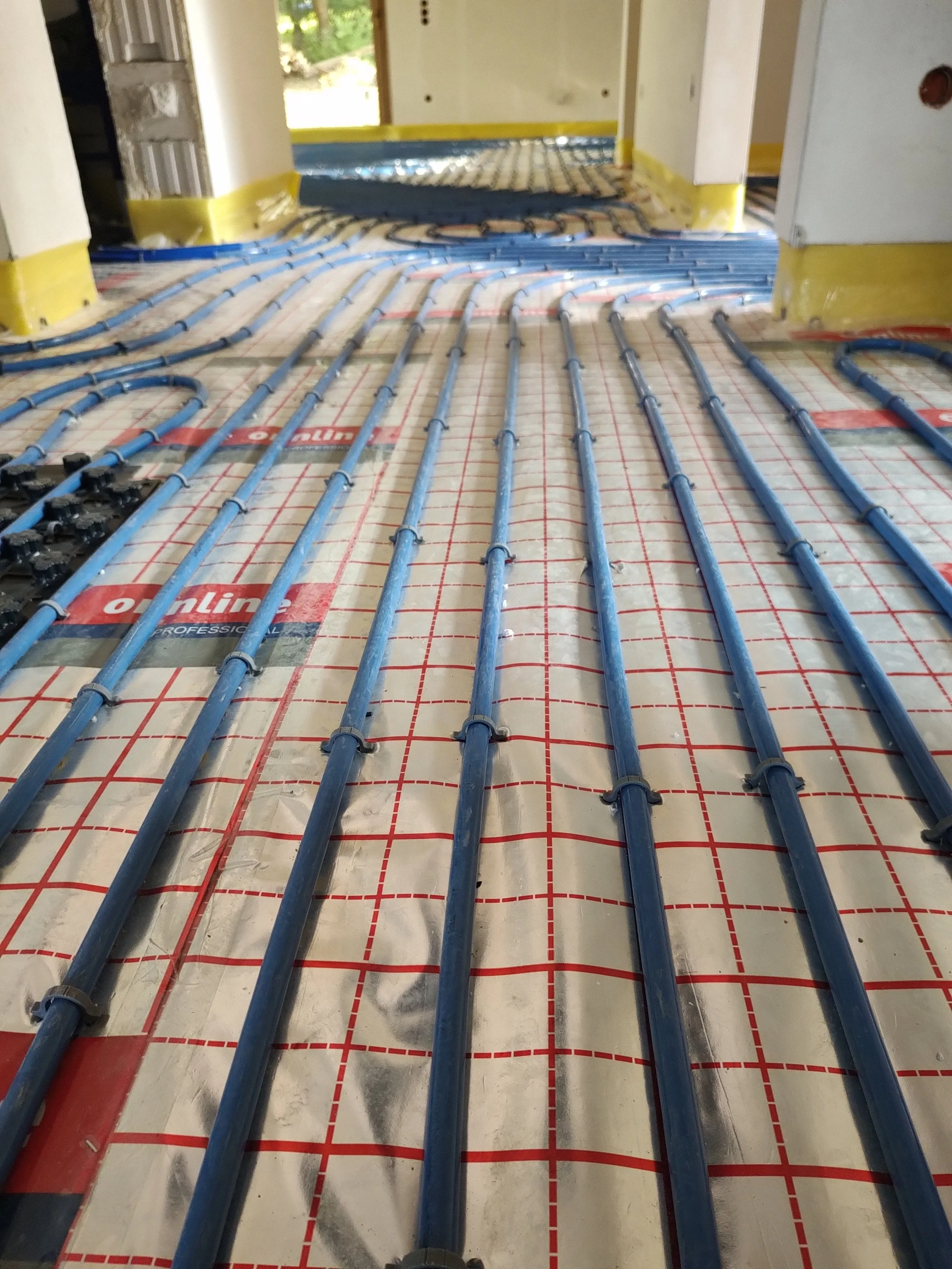 Underfloor heating pipes being installed in a building's floor, with blue PEX pipes laid out on a red and white grid system, ready to be embedded in concrete.