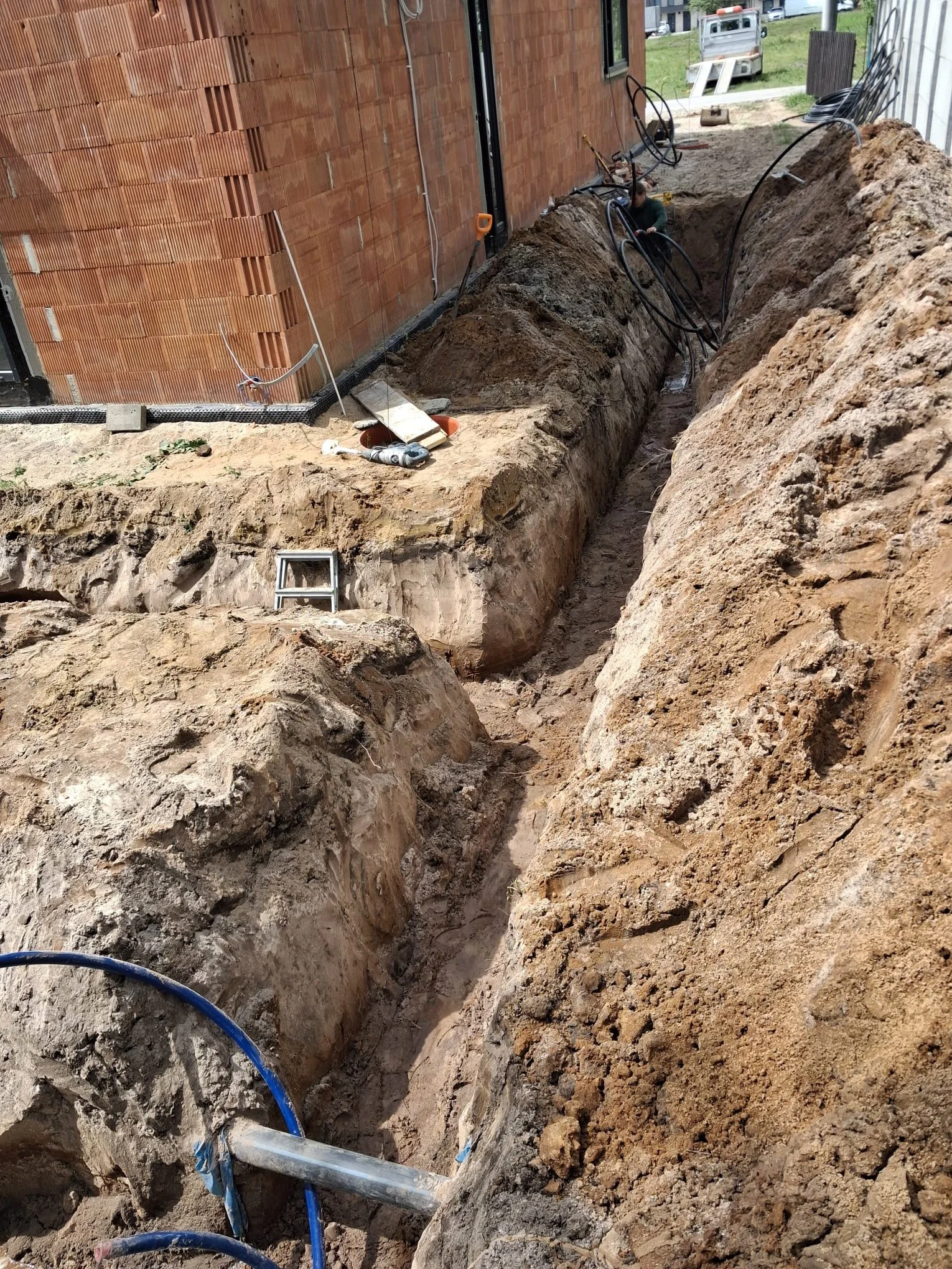 Construction site with a large trench dug alongside a brick building, exposed dirt, and construction tools and pipes visible.