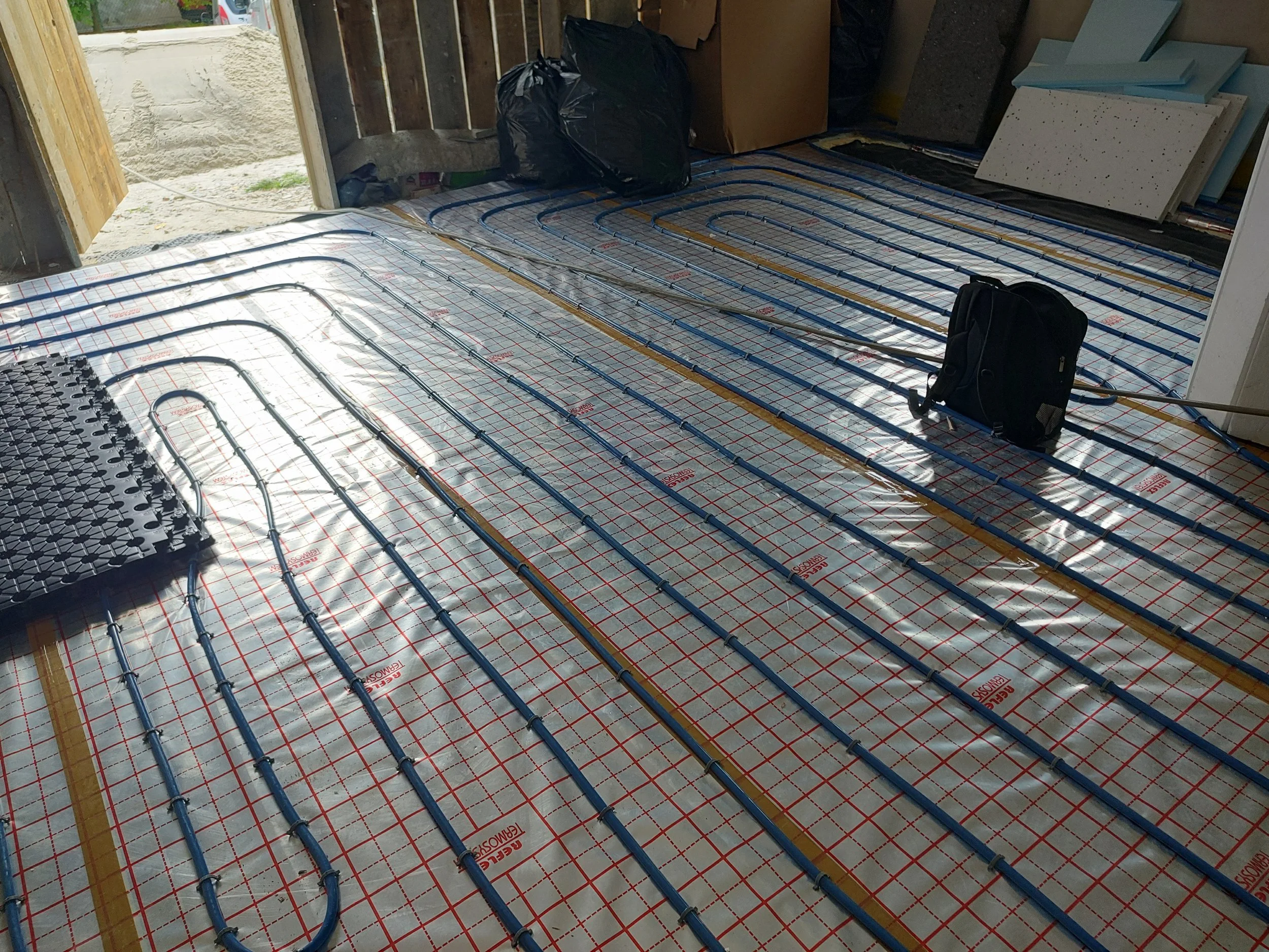 Installation of radiant floor heating pipes on a construction floor with insulation and grid, in a partially built room.