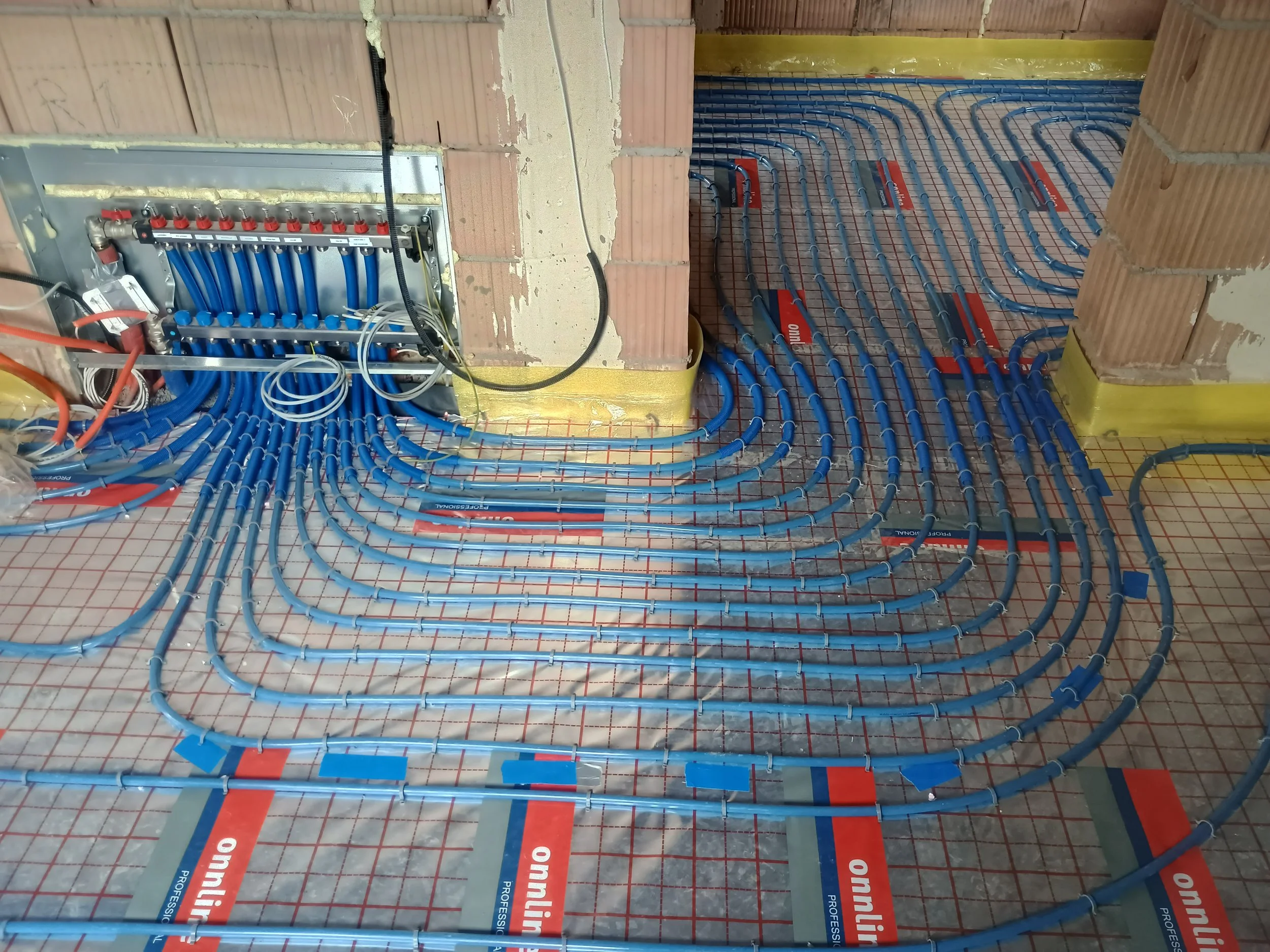 Under construction floor with red and blue plastic pipes arranged in a pattern, a metal electrical box with red and blue valve handles, and part of a brick wall.