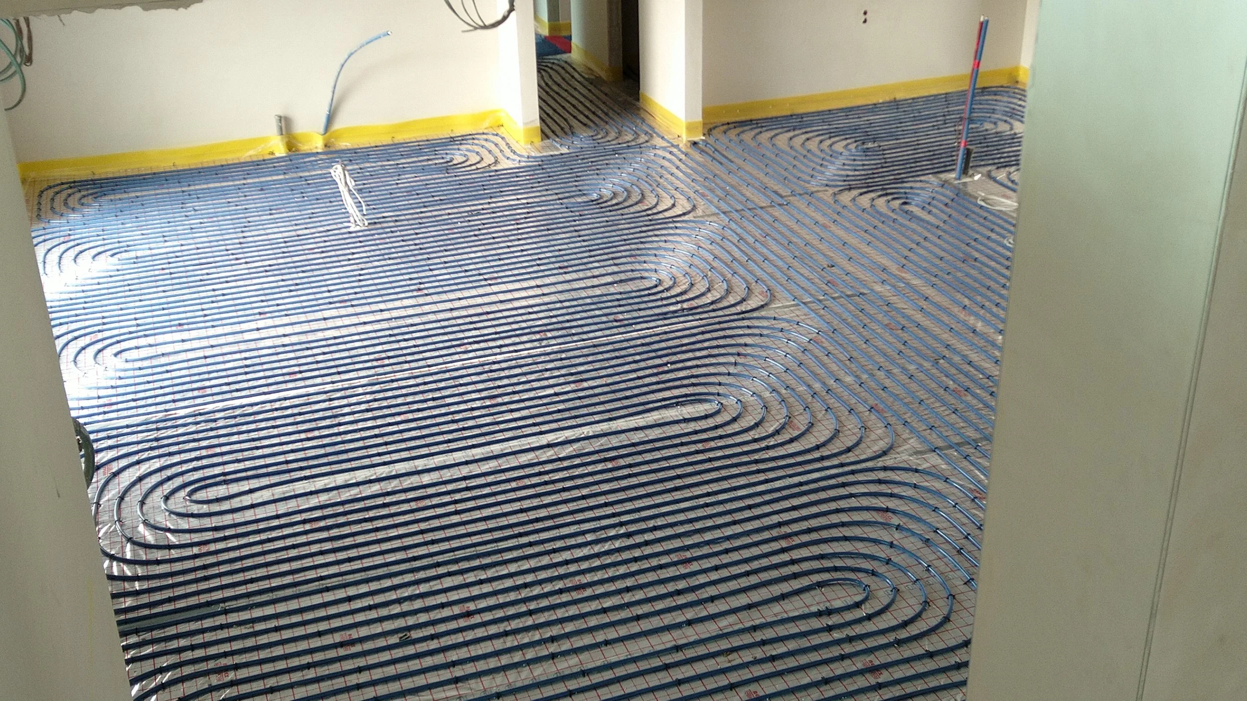 Underfloor heating pipes installed in a room, with yellow masking tape along the edges of the floor. The pipes are embedded in a reflective insulation layer, and there are electrical wires protruding from the wall.