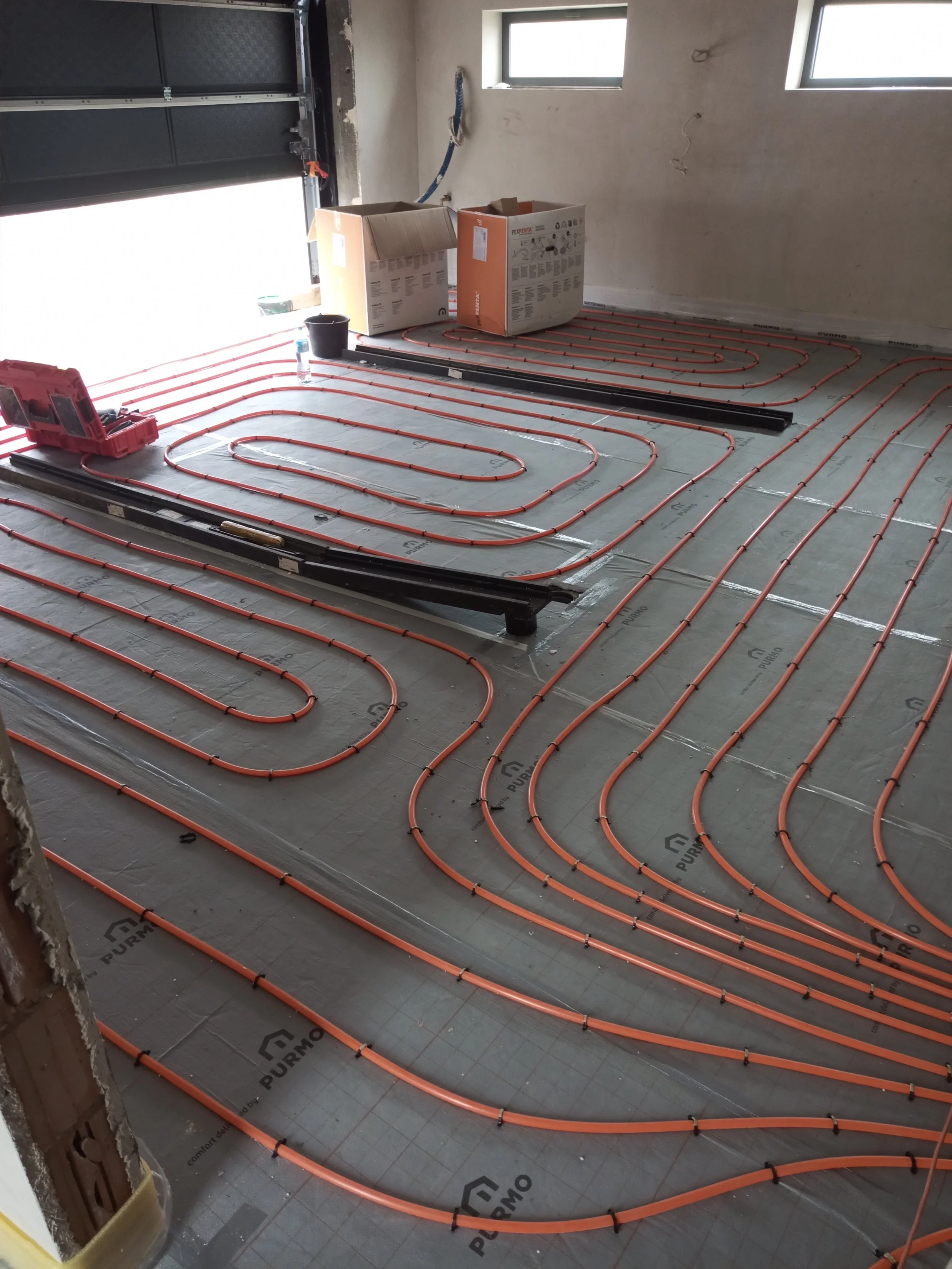 Under construction room with orange tubing for radiant floor heating laid out on the concrete floor, boxes, and a black pipe or railing at the back.