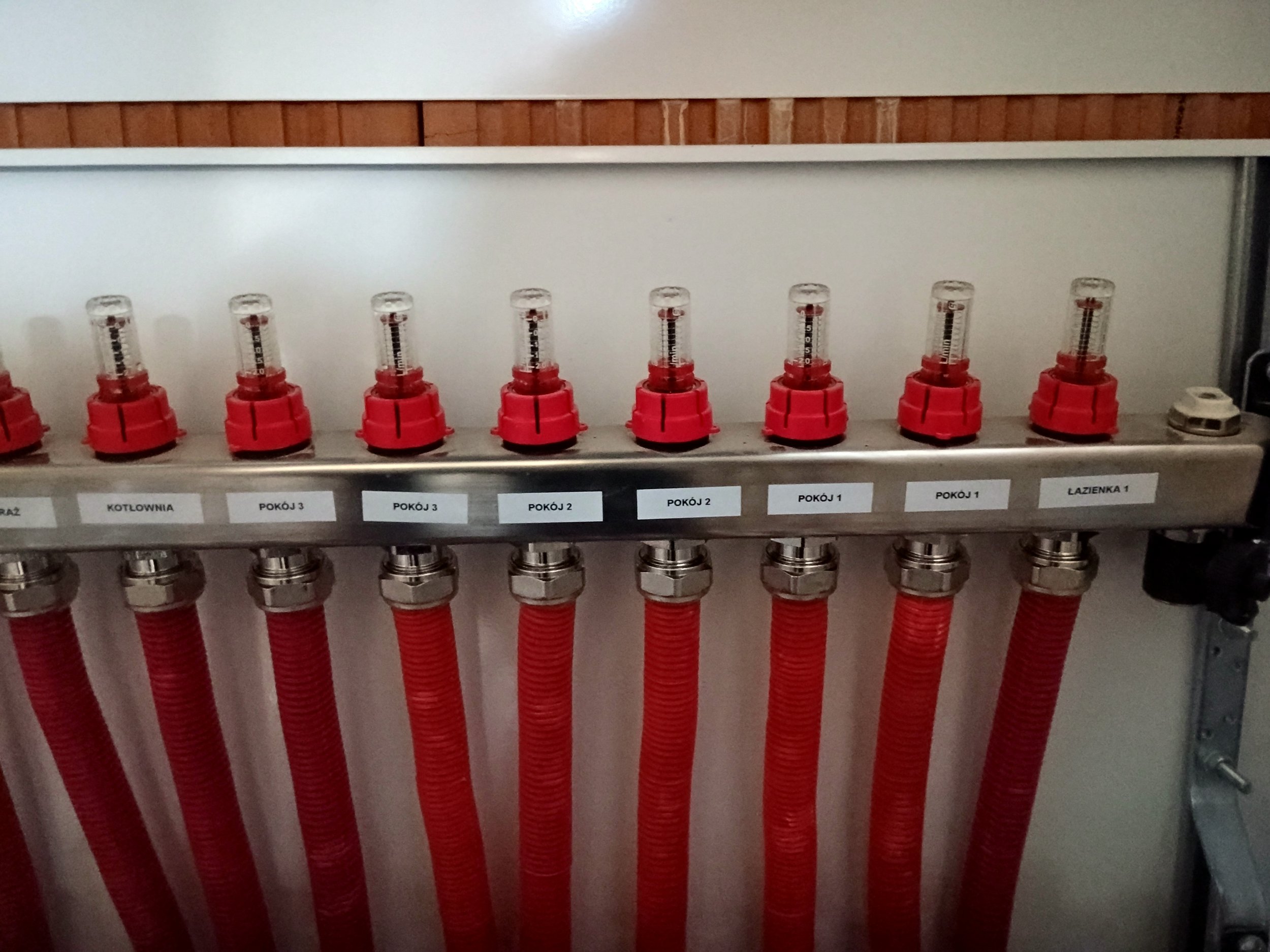 A row of red plastic pipes and clear measuring devices mounted on a metallic panel, with small labels below in Polish.