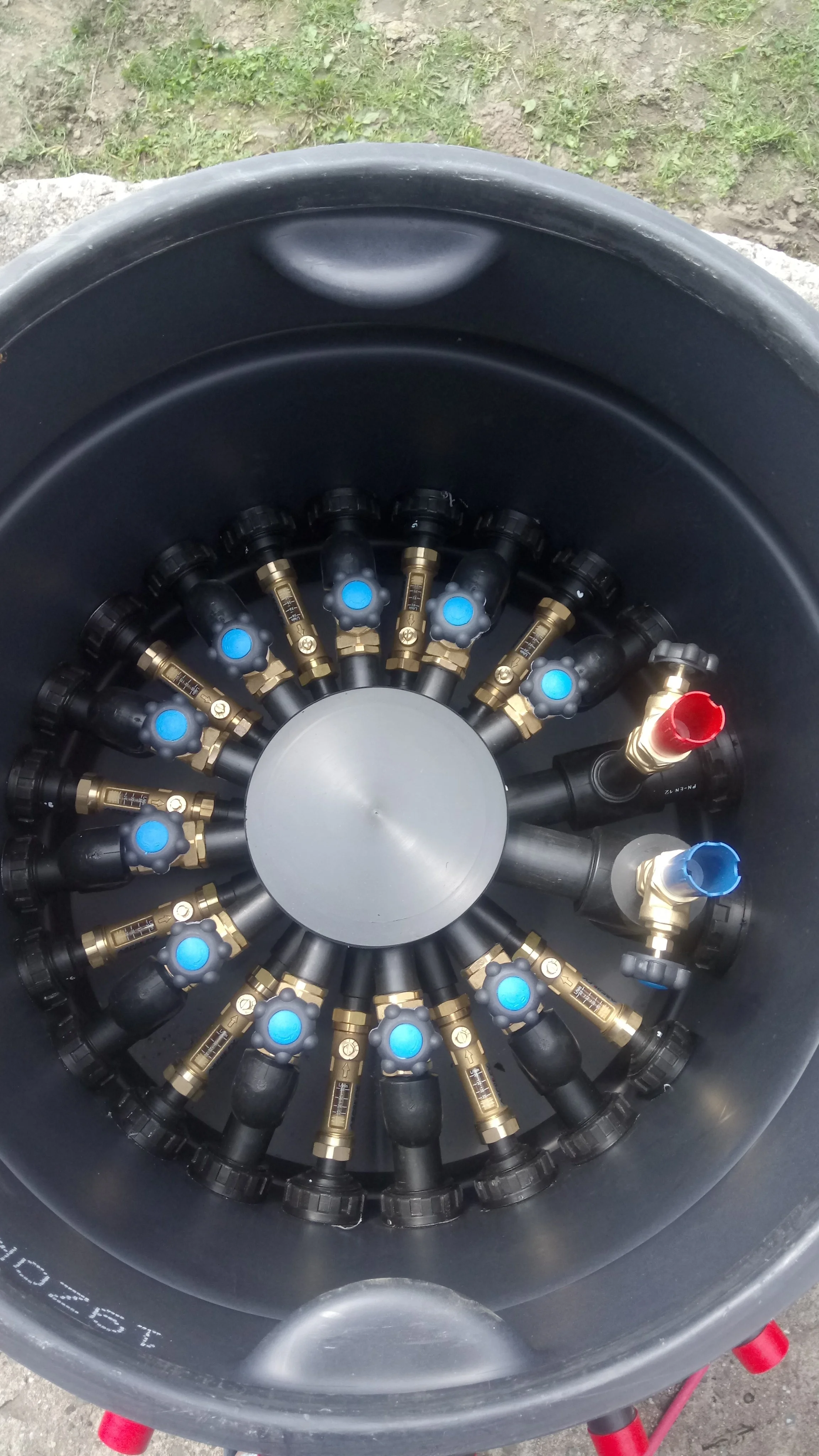 Image of a circular manifold with multiple brass and black connectors, some with blue and red caps, arranged in a radial pattern inside a black cylindrical container.