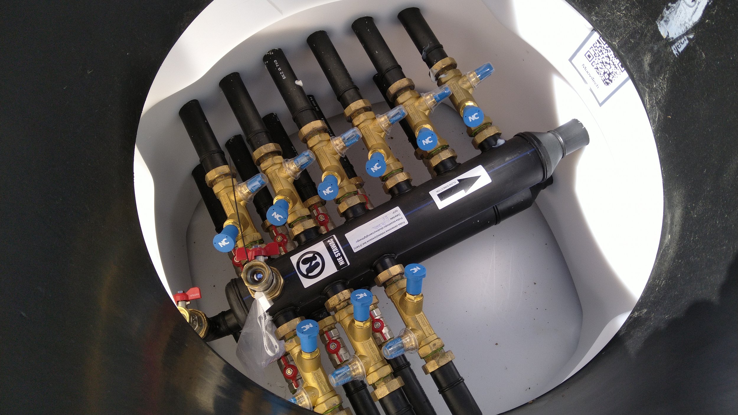 Multiple black pipes connected with gold-colored valves and blue and red circular indicators inside a white cylindrical tank.
