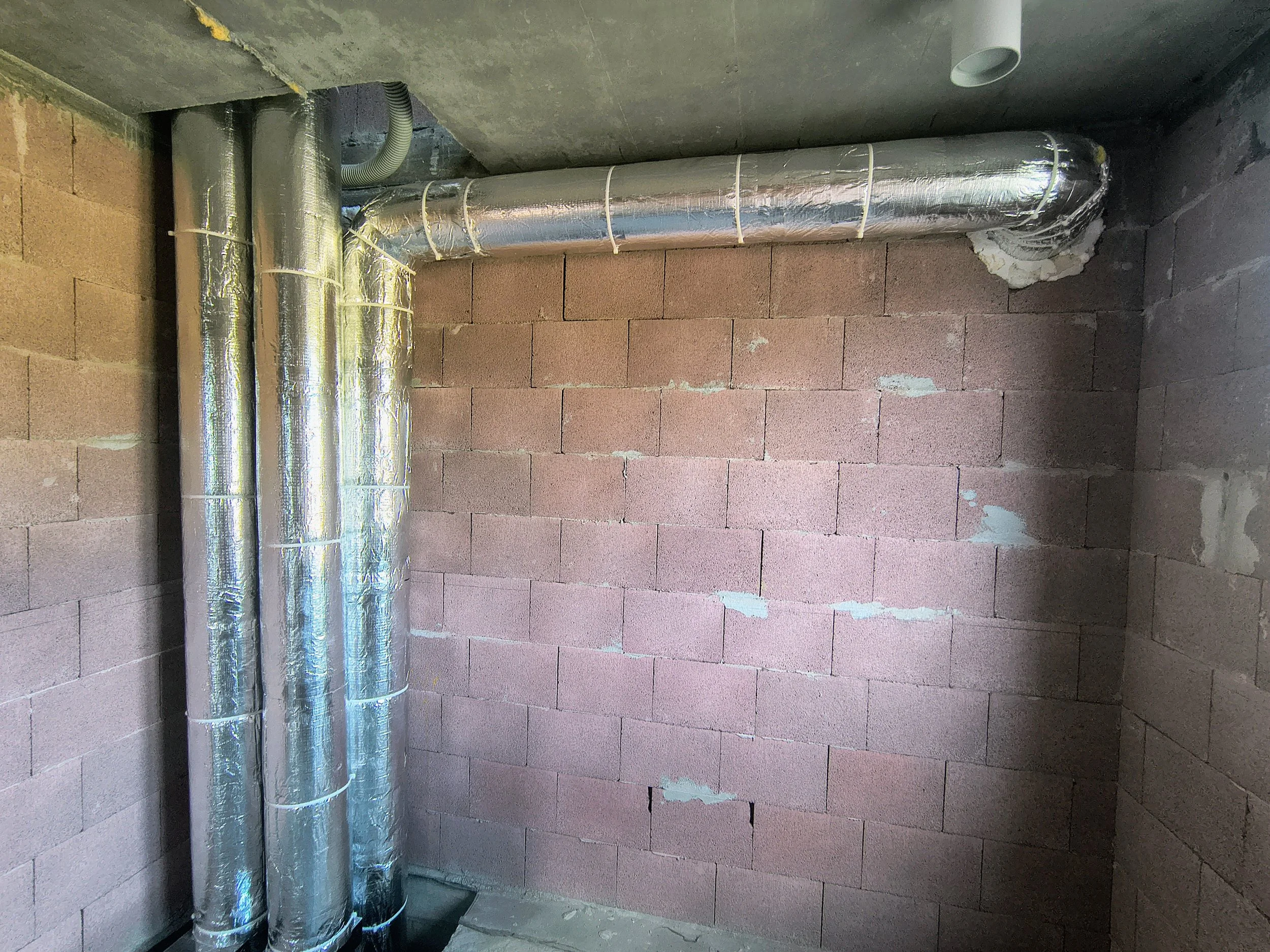 Four silver insulated pipes run vertically along a brick wall in a basement or utility room. The pipes are secured with white straps and are connected at the top to an elbow joint. The wall is unfinished with visible mortar and some patches of white 
