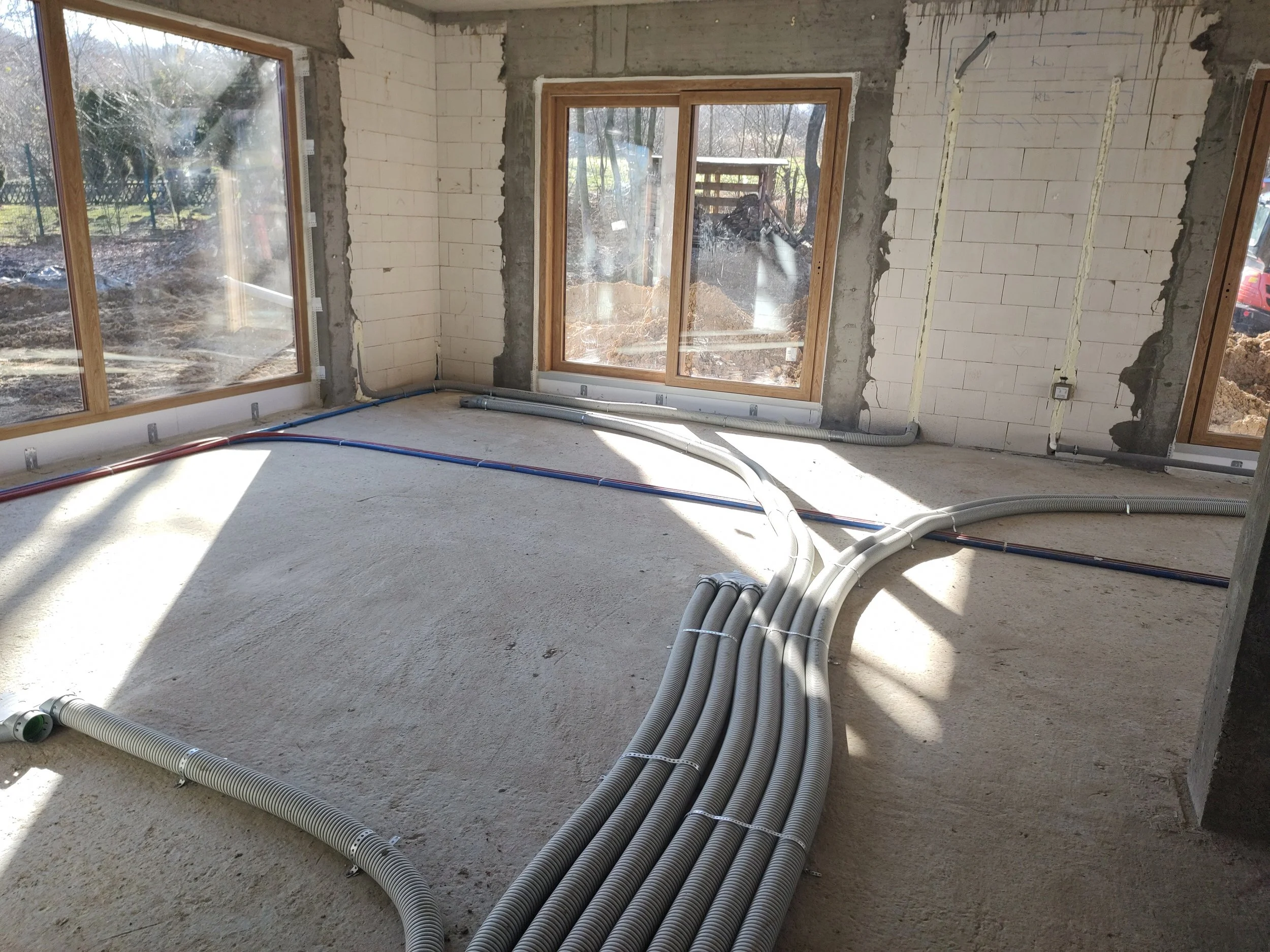 Interior of a house under construction featuring exposed concrete walls, large windows, and electrical conduits running across the unfinished floor.