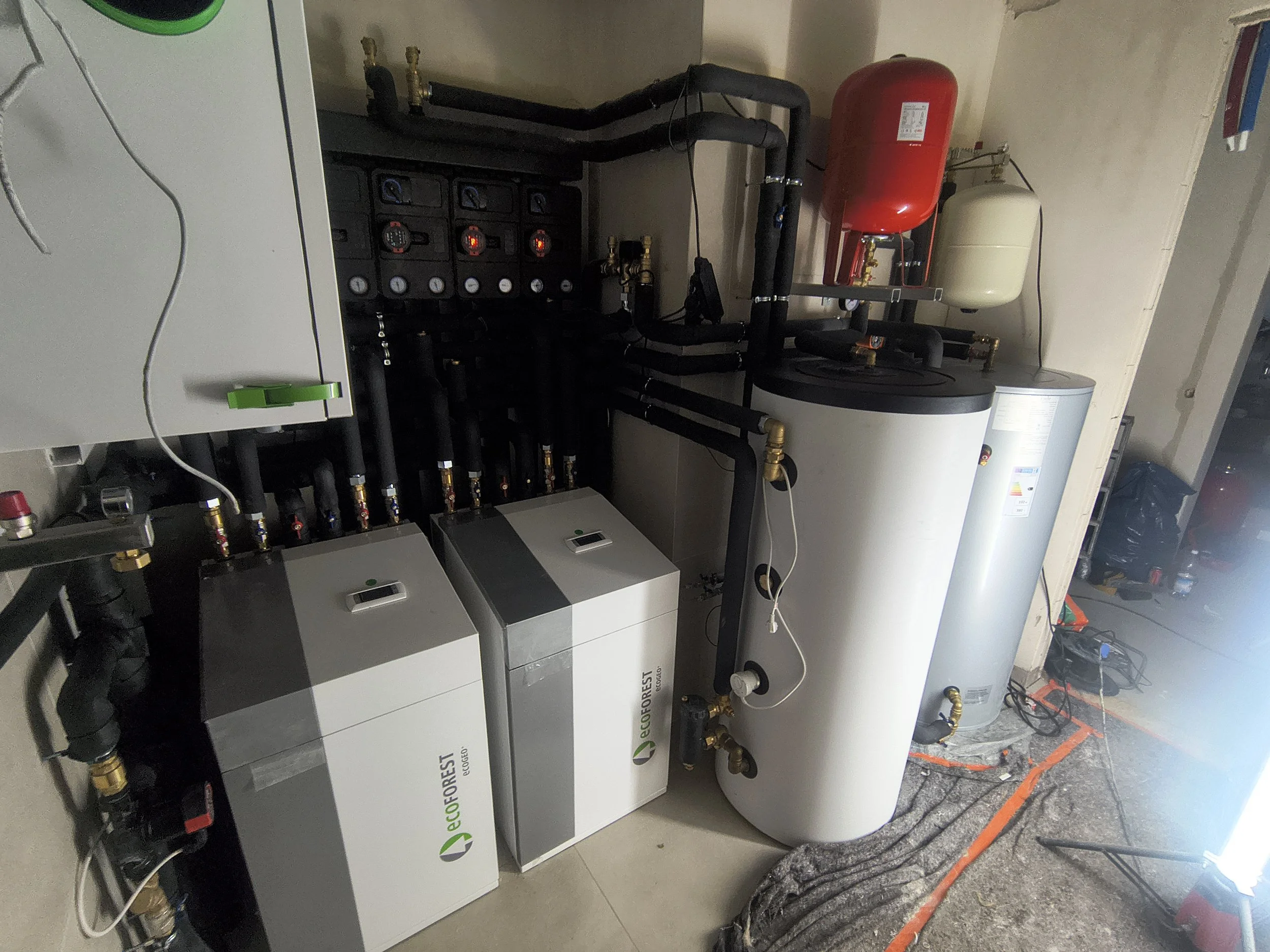 A mechanical room features a water heater, red expansion tank, and a system of pipes and gauges. The equipment is labeled with the Ecoforest logo.