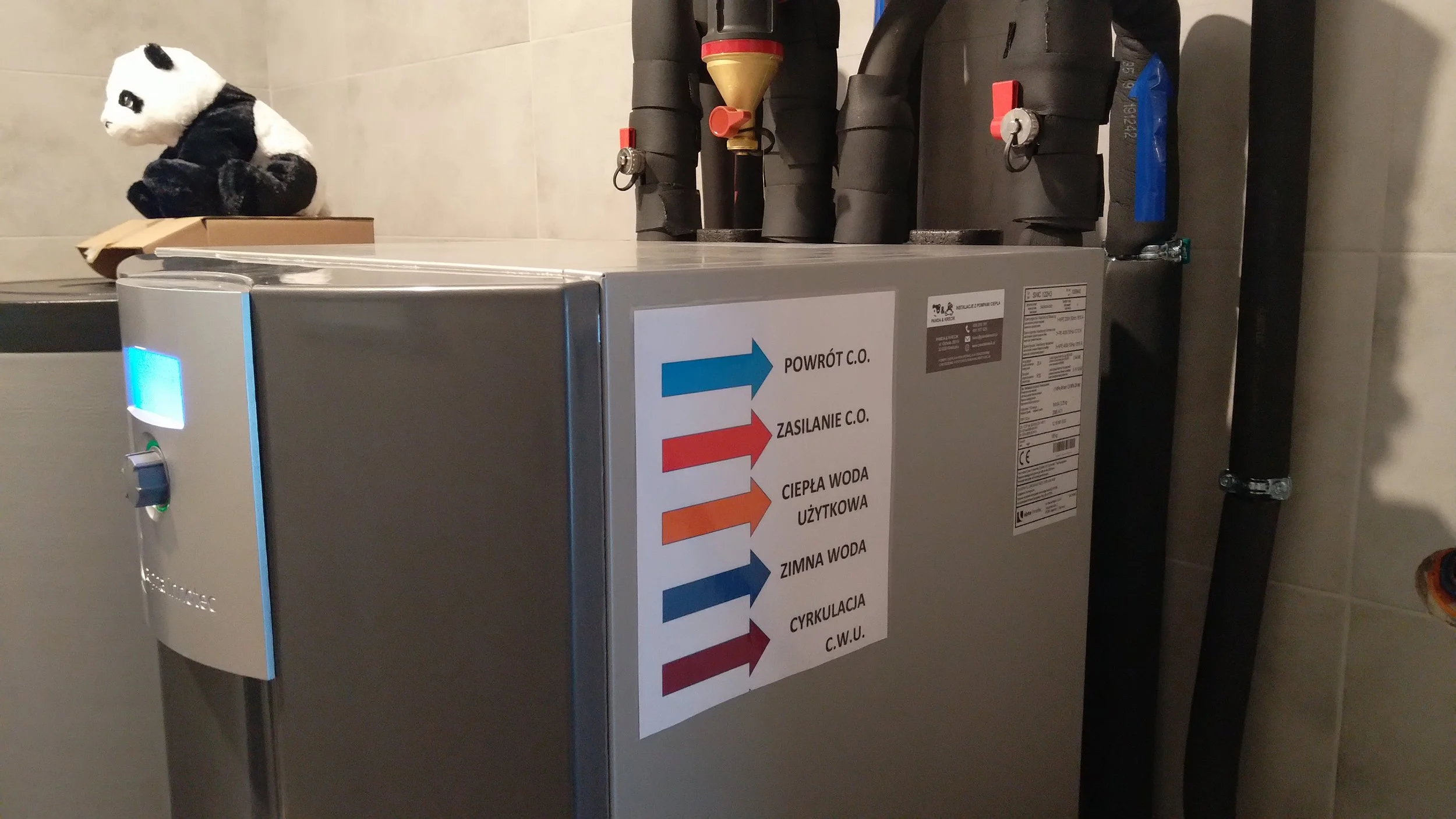 A domestic boiler unit with pipes and a colorful label showing various Polish terms for water and heating systems, including directions for turning on and off.