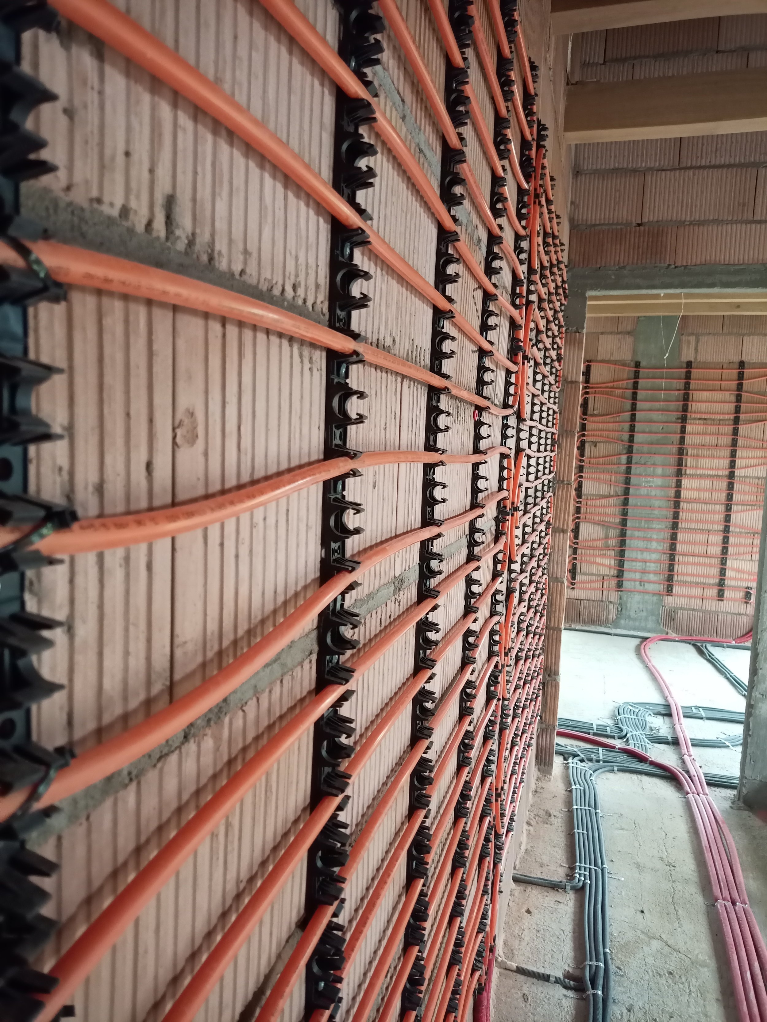 Rows of orange pipes and black brackets installed on a wooden wall at a construction site, with additional pink and blue pipes running along the floor and wall.