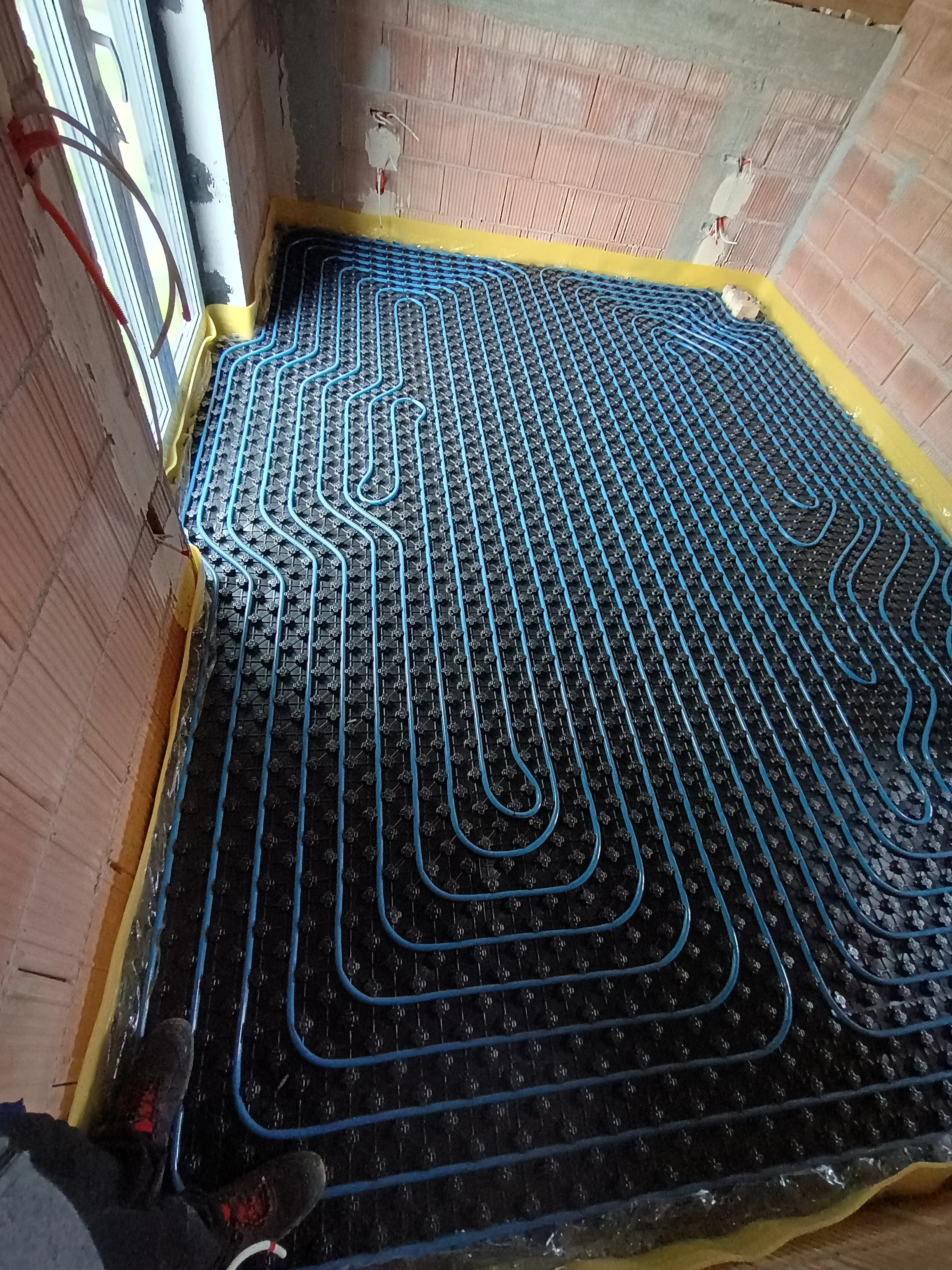 Installation of underfloor radiant heating system with blue pipes laid in a serpentine pattern on a black insulation panel inside a room under construction, with brick walls and a window.