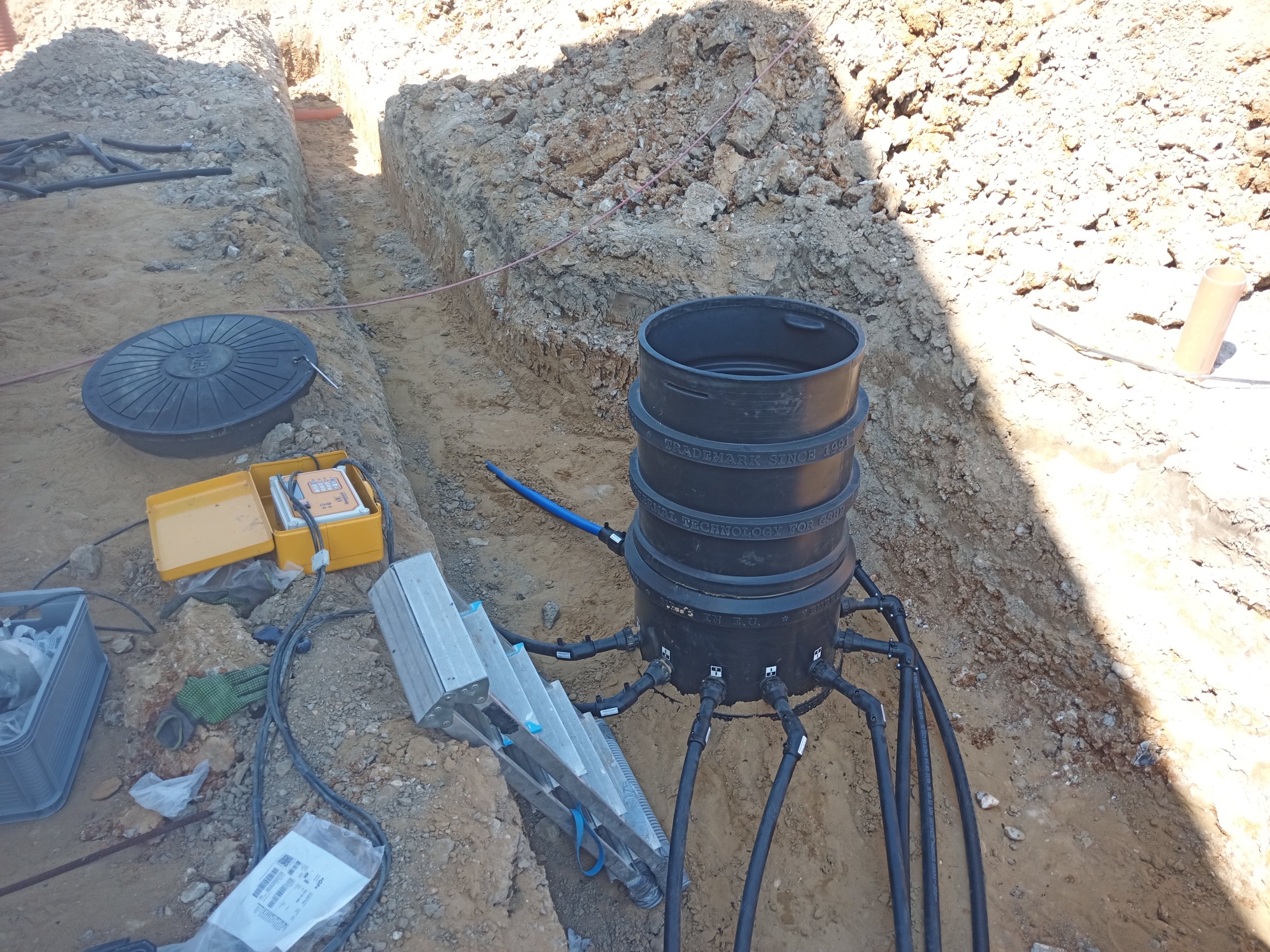 A construction site showing a deep trench with disturbed soil and rocks. There are black pipes, a black cylindrical object, a yellow toolbox, various tools and cables scattered around, and dirt and rocks on the ground.