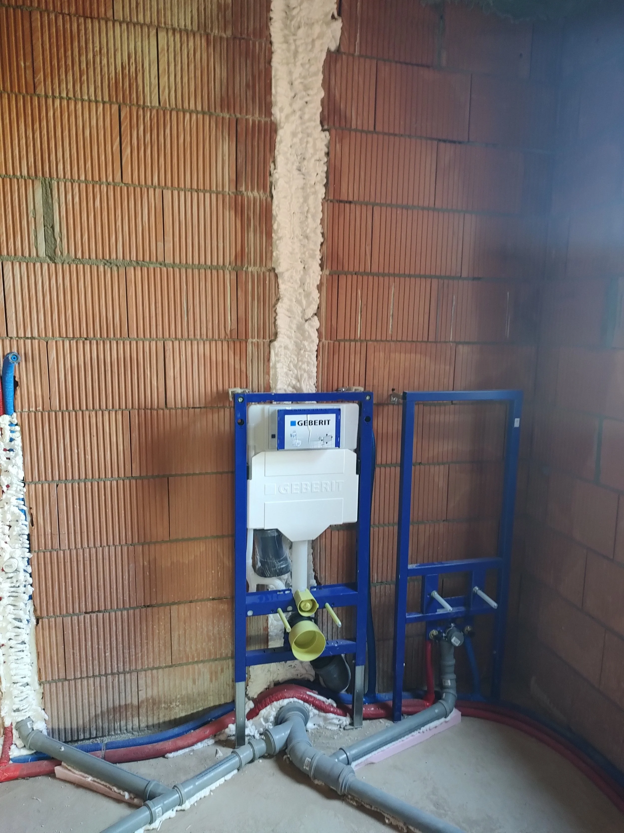 Construction site with exposed brick wall and plumbing pipes, with a Geberit toilet frame installed but not yet finished.