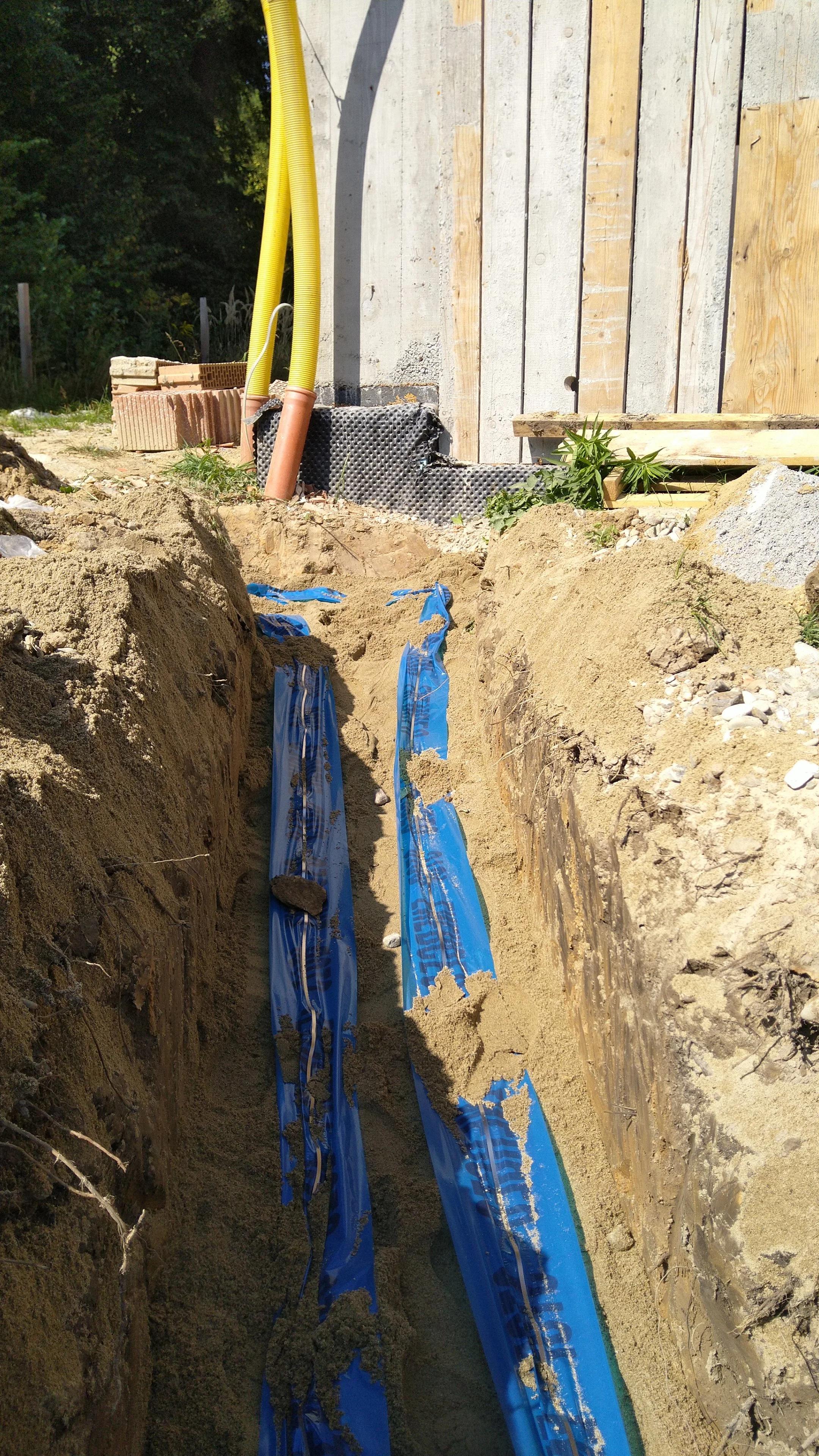 A construction site showing a trench with blue waterproofing membrane, pipes, and sand, next to a partially built wooden structure with yellow electrical conduit pipes.