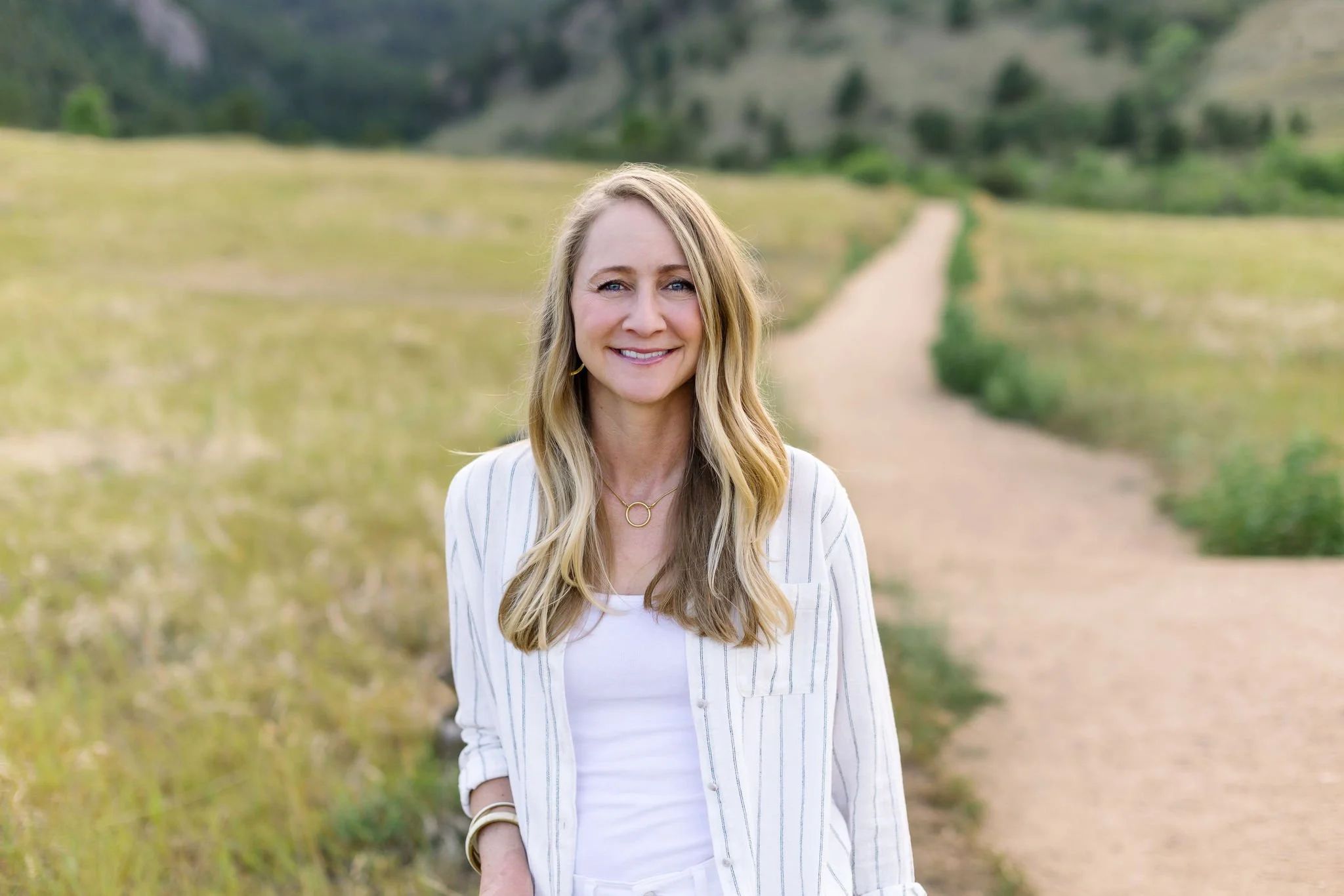 A woman with long blonde hair smiling outdoors on a trail with greenery and hills in the background.