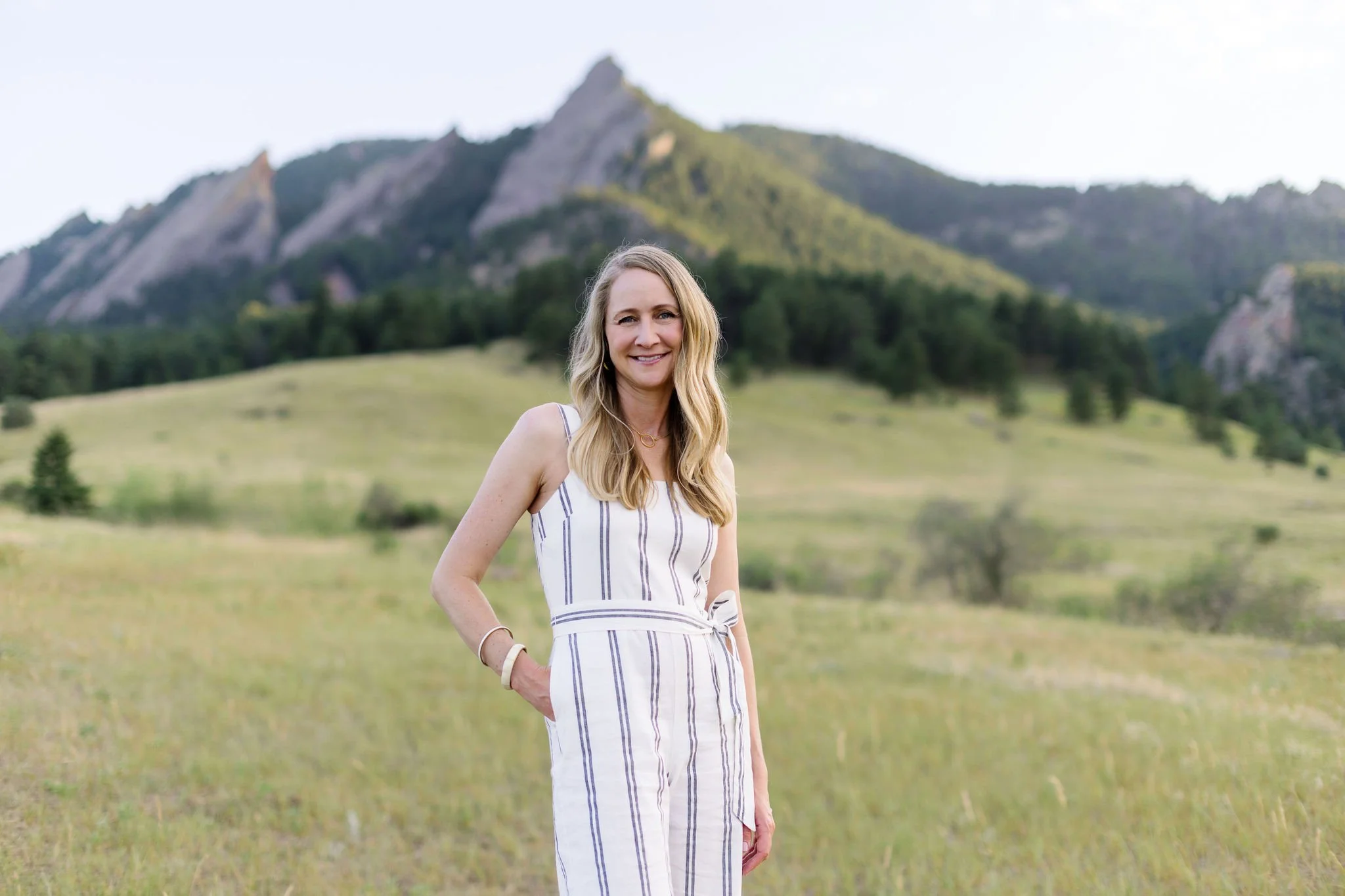A woman with long blonde hair smiling and standing in a grassy field with a mountain range in the background.