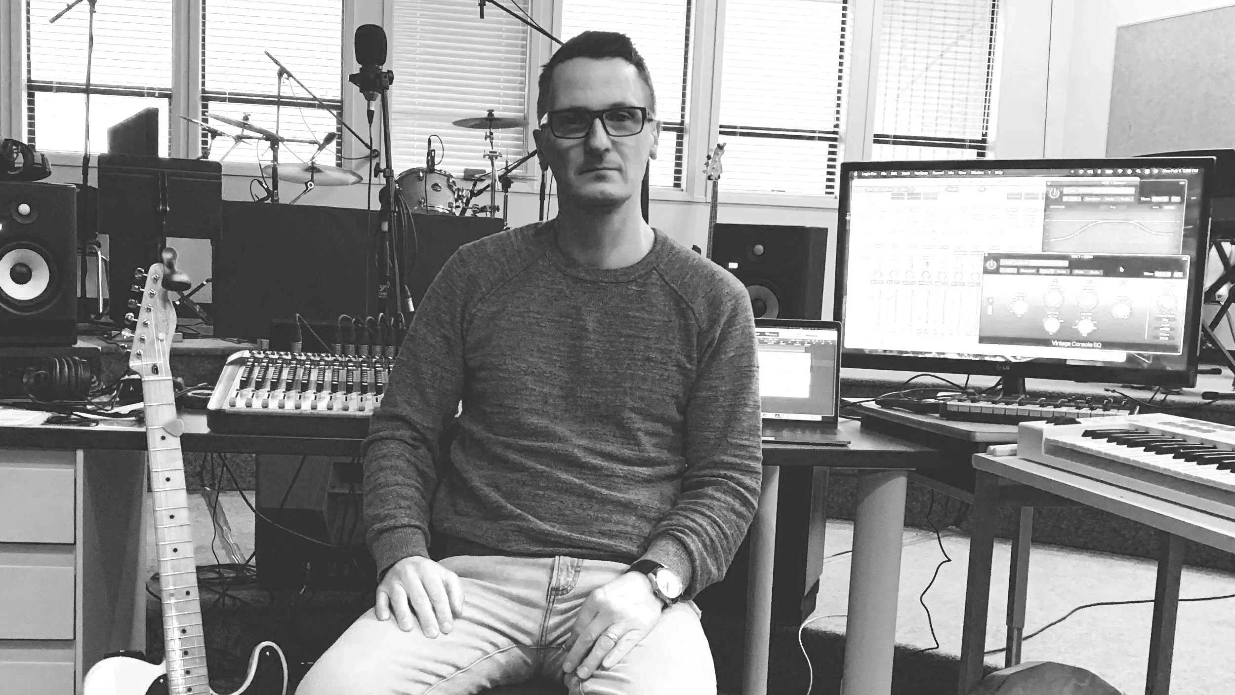 A-man-wearing-glasses-sitting-in-a-music-studio-with-musical-instruments-and-recording-equipment-around-him