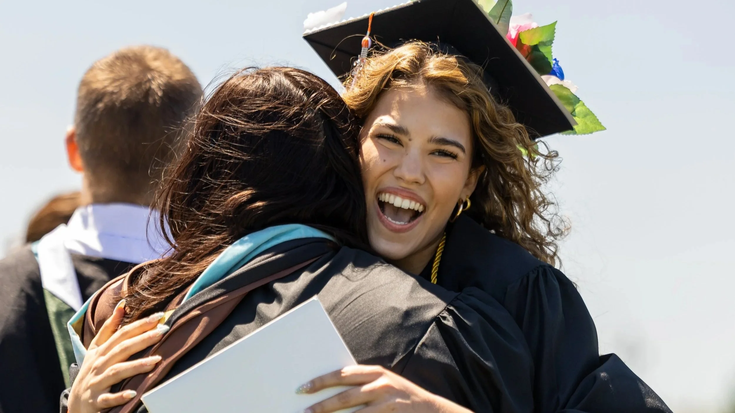 A young woman in a graduation cap and gown hugging someone at her graduation ceremony, smiling joyfully.