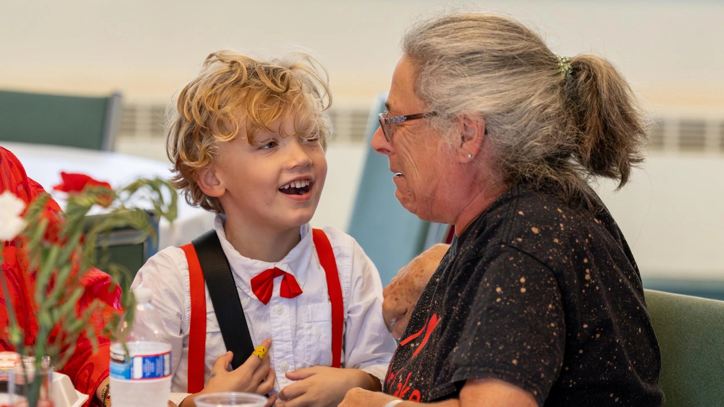 A young boy with curly blonde hair and a red bow tie is talking to an older woman with gray hair tied back, glasses, and earrings, in a room with a table and some items including a water bottle and flowers.