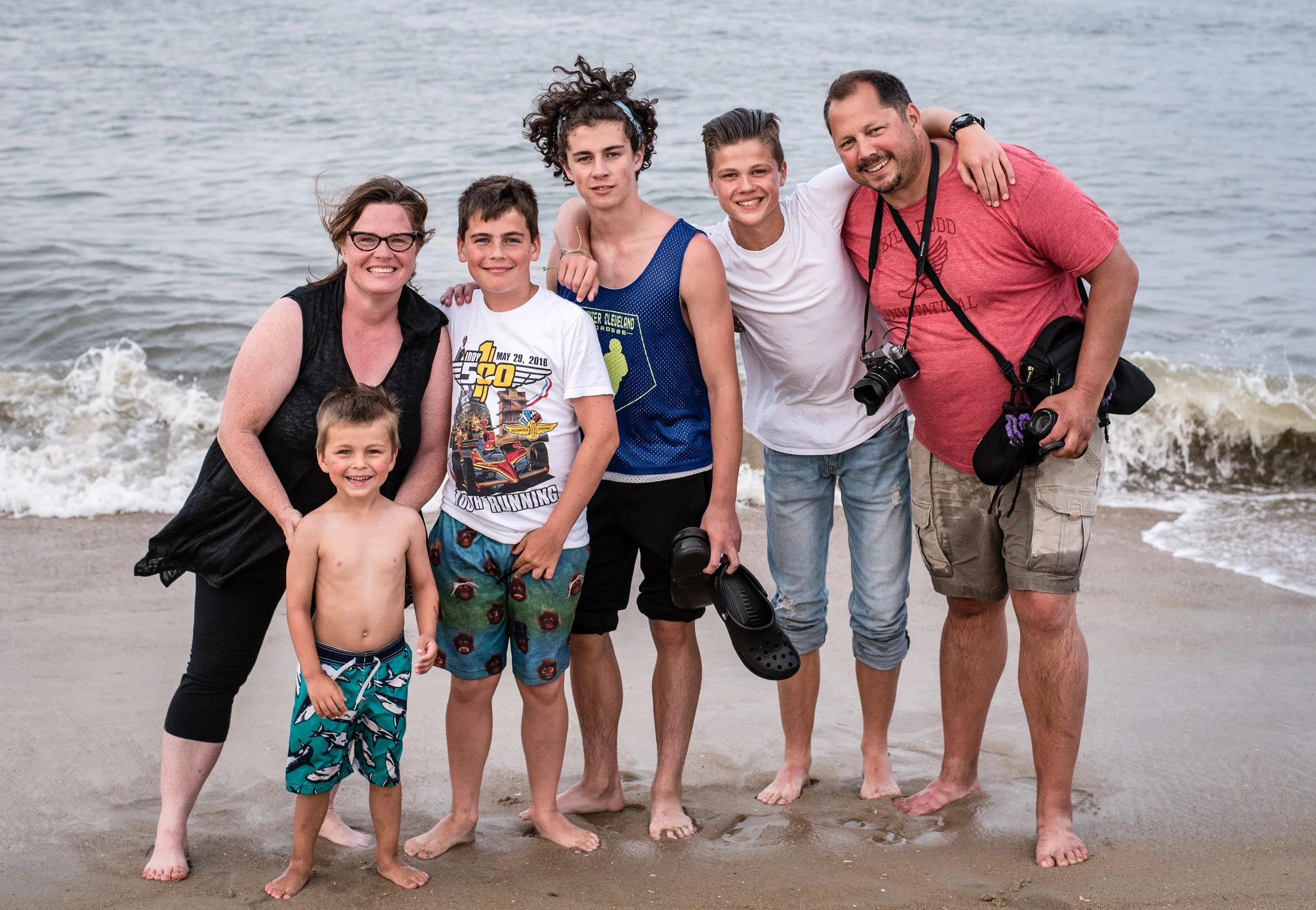 A group of seven people, including two adults and five children, standing on a sandy beach by the water, smiling and enjoying a day at the seaside.
