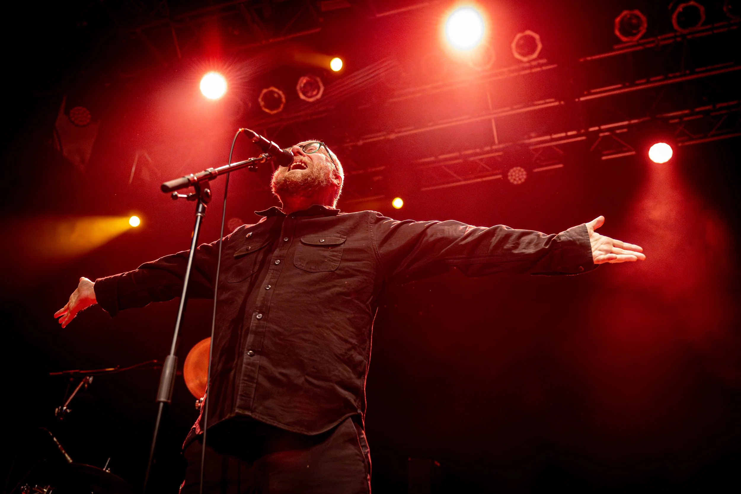 A man with glasses and a beard singing passionately at a microphone on stage with red and yellow stage lights overhead, arms outstretched.