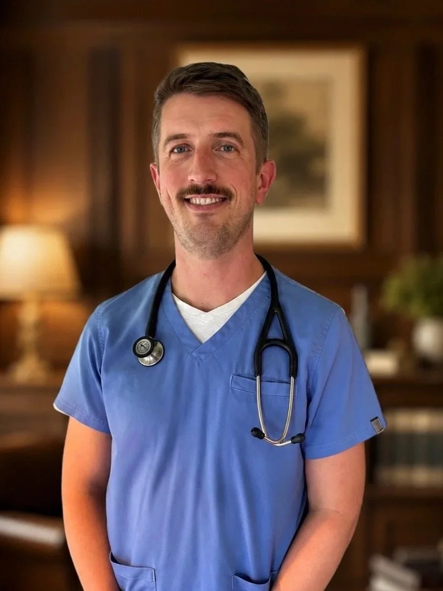 A young male healthcare professional in blue scrubs with a stethoscope around his neck, smiling in a warmly lit room with wooden paneling and artwork in the background.