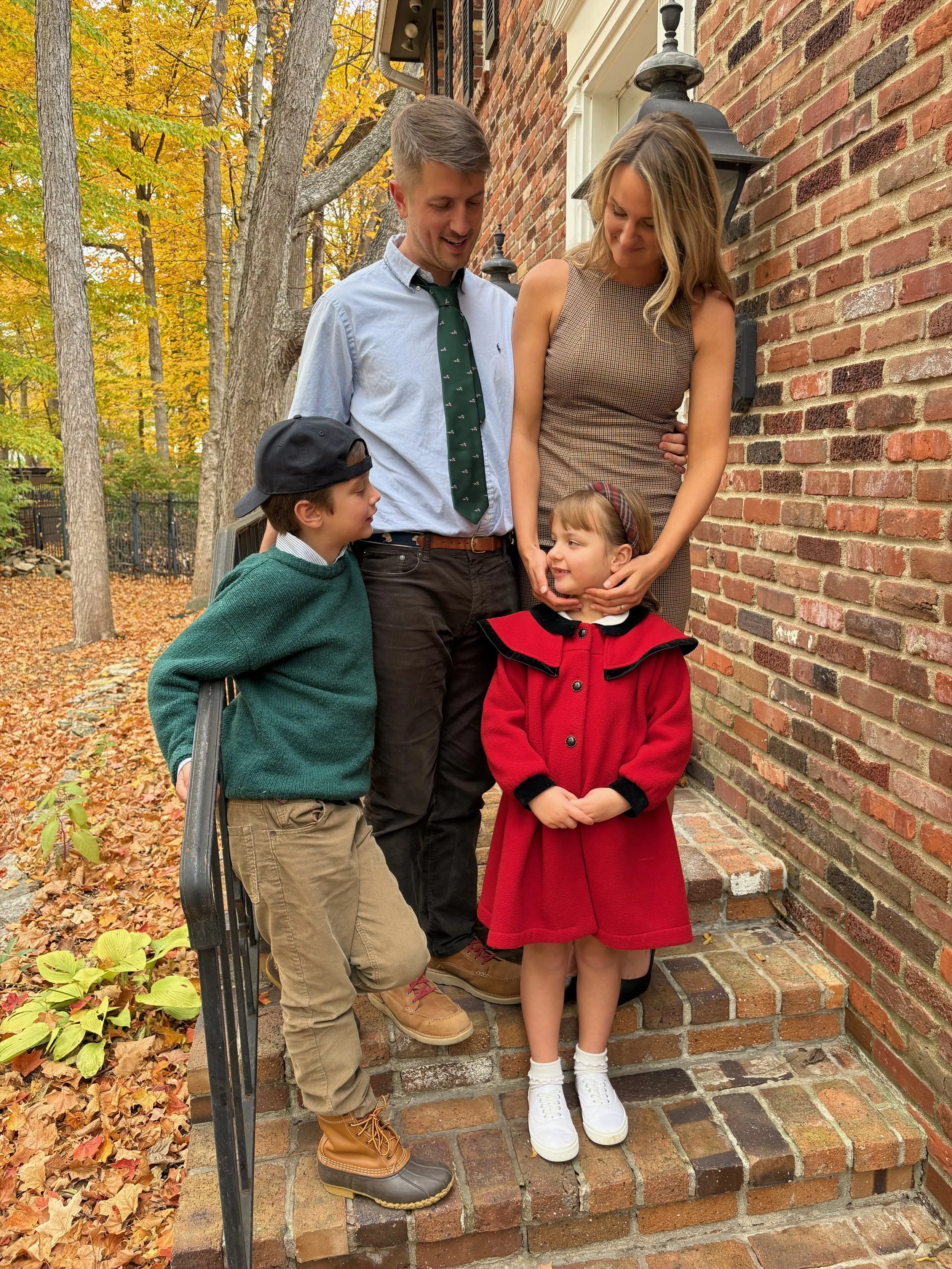 A family of four, including a man, a woman, and two children, standing on brick steps outside a brick house with fall foliage in the background. The man and woman look lovingly at their children.