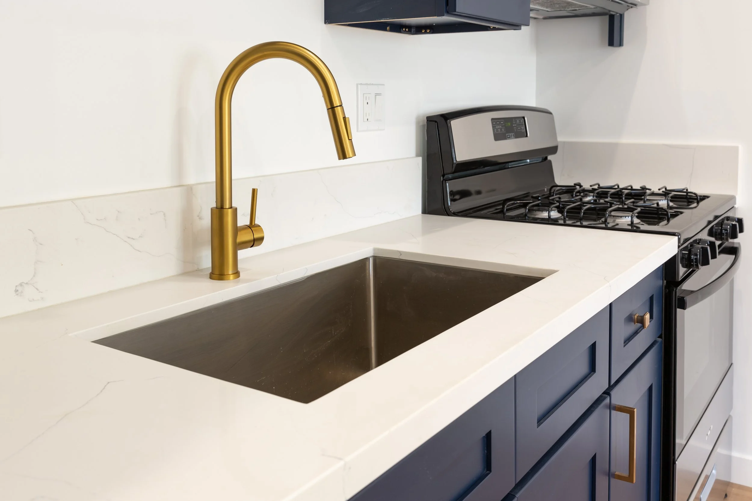 Kitchen countertop with white marble surface, gold faucet at sink, black stove, and dark blue cabinets.