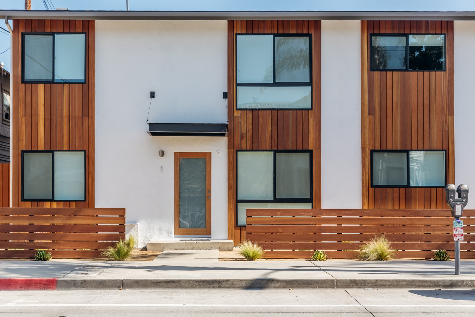 Modern two-story residential building with a wooden and white facade, multiple large windows, a wooden fence in front, and a parking meter on the sidewalk.